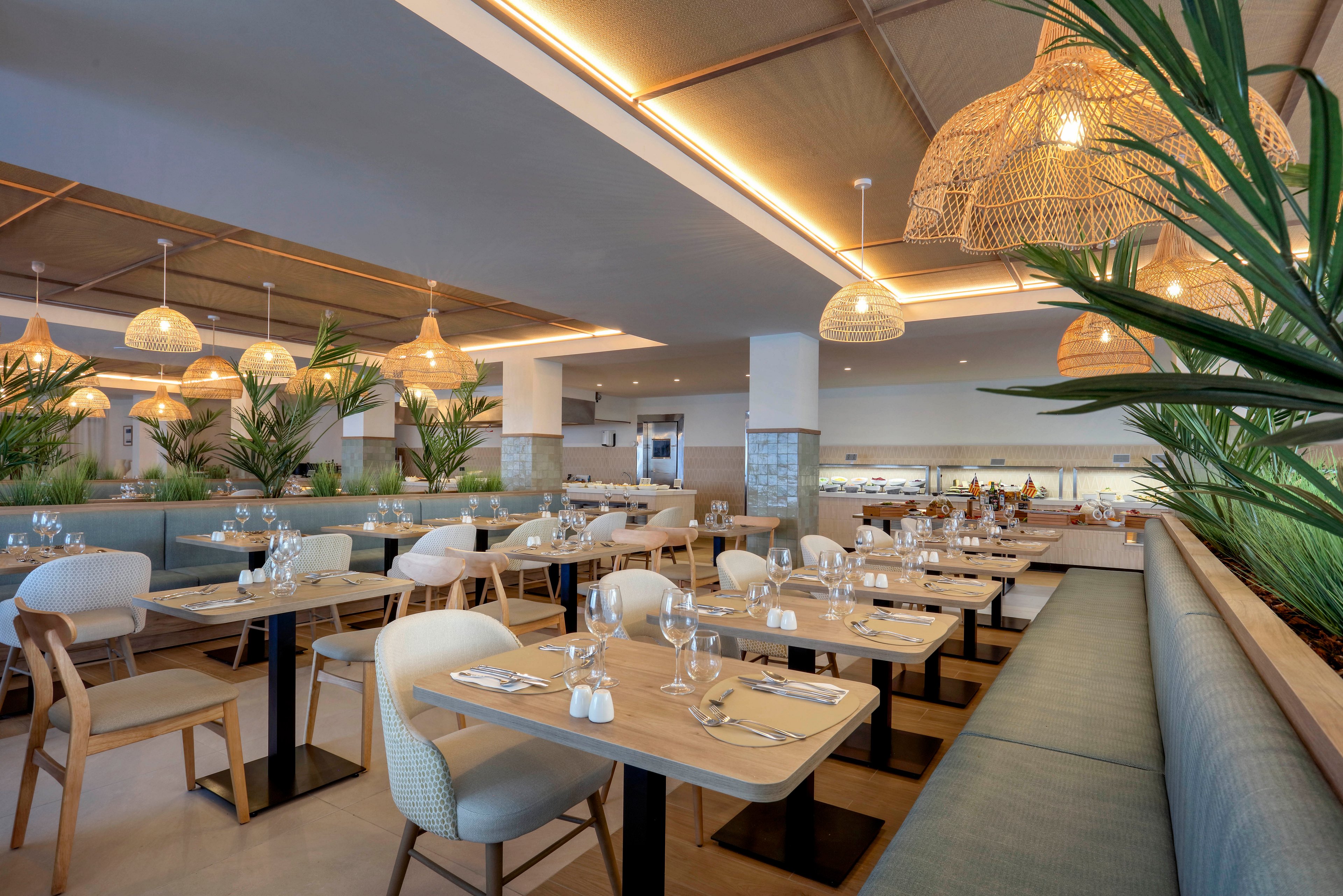 A contemporary restaurant dining area features neatly arranged wooden tables and cushioned chairs. Tables are fully set with wine glasses, water glasses, plates, cutlery, and small salt and pepper shakers. Large green plants and woven rattan pendant lights create a relaxed, coastal atmosphere. Soft ceiling lighting and pale blue booth seating add to the bright, inviting interior design.