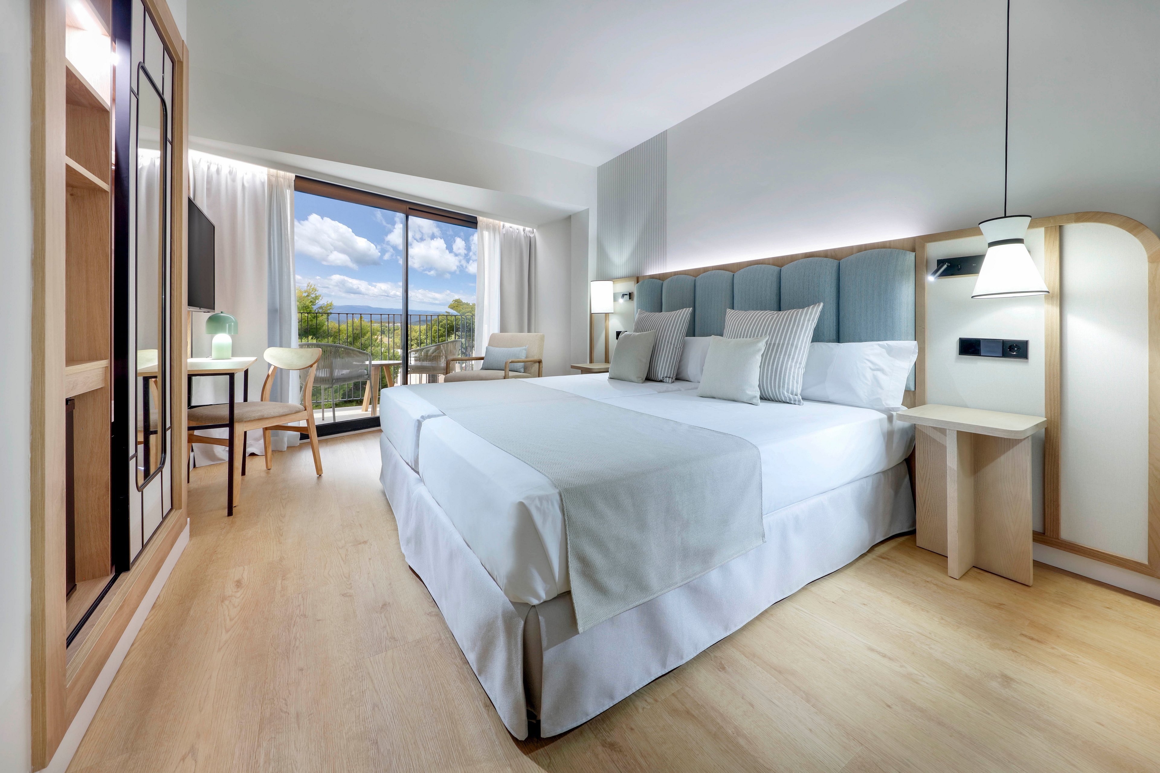 A spacious, contemporary hotel room features a large neatly made bed with white linens and soft blue upholstered headboard. Light wood flooring and minimalist furniture create an airy, clean atmosphere. A desk and chair sit near a large glass door that opens to a balcony with views of greenery and a partly cloudy sky. Soft natural light fills the room, complemented by modern pendant and bedside lighting.