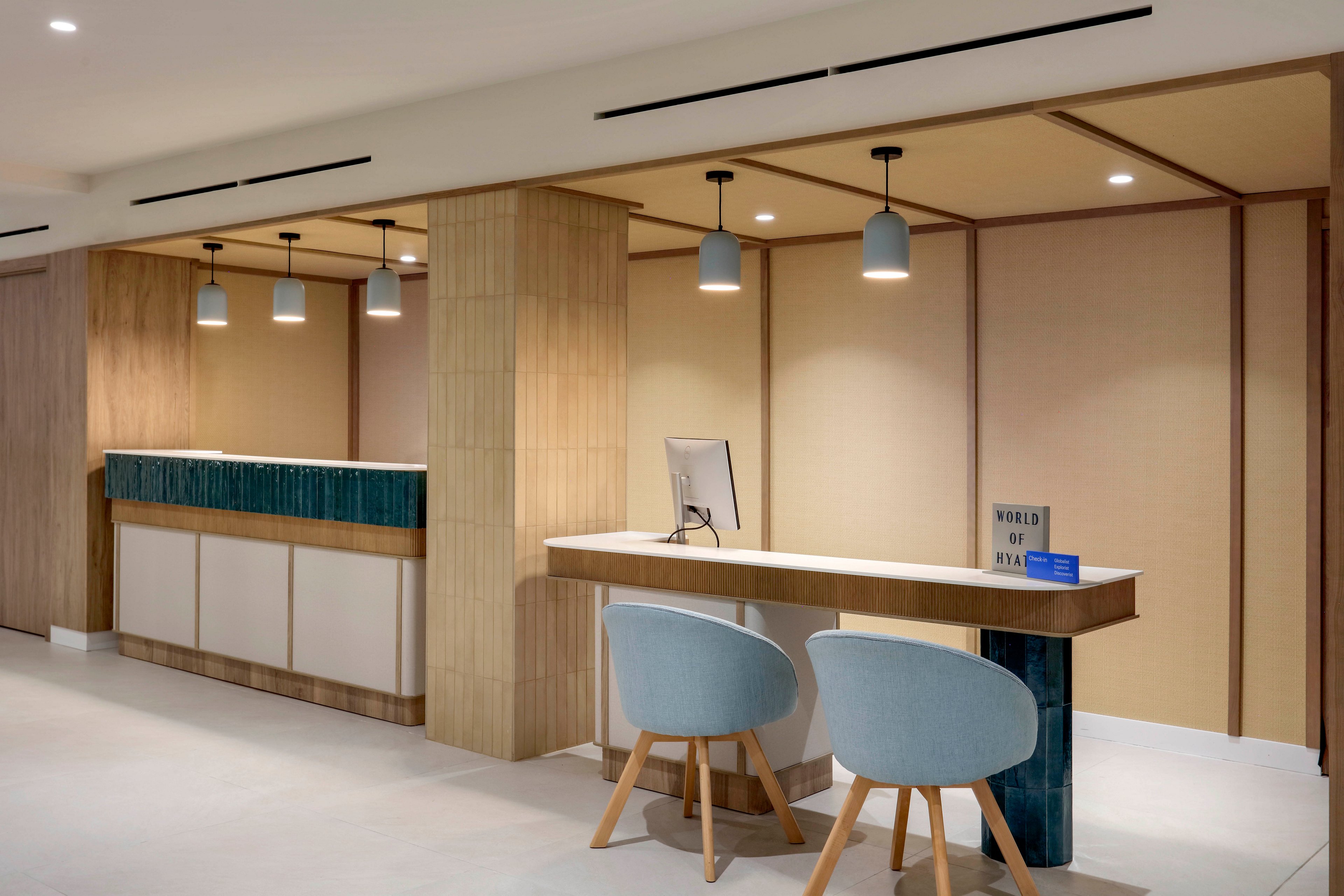 Contemporary hotel reception area featuring a main check-in counter and a smaller desk with two upholstered chairs. The space is clean and minimalist, with light wood finishes and teal accent panels. Pendant lights hang from the ceiling, illuminating the workstations and computer monitor. A small sign on the desk reads "WORLD OF HYATT," indicating the hotel affiliation.