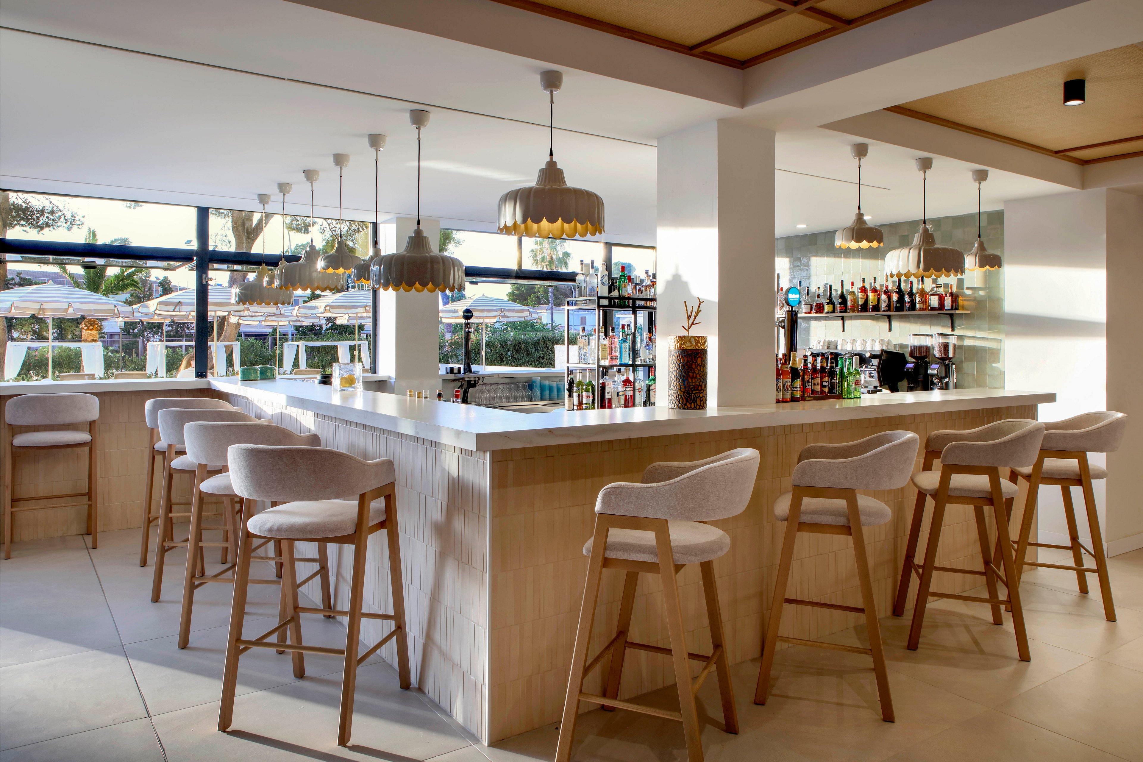 A contemporary bar interior features a light wood counter surrounded by cushioned wooden bar stools. Pendant lights hang above the bar, illuminating shelves stocked with various bottles and glassware. Large windows open to an outdoor terrace with sun loungers and umbrellas, suggesting a resort or vacation setting. The overall design is bright and minimal, with neutral tones and clean lines creating a relaxed atmosphere.
