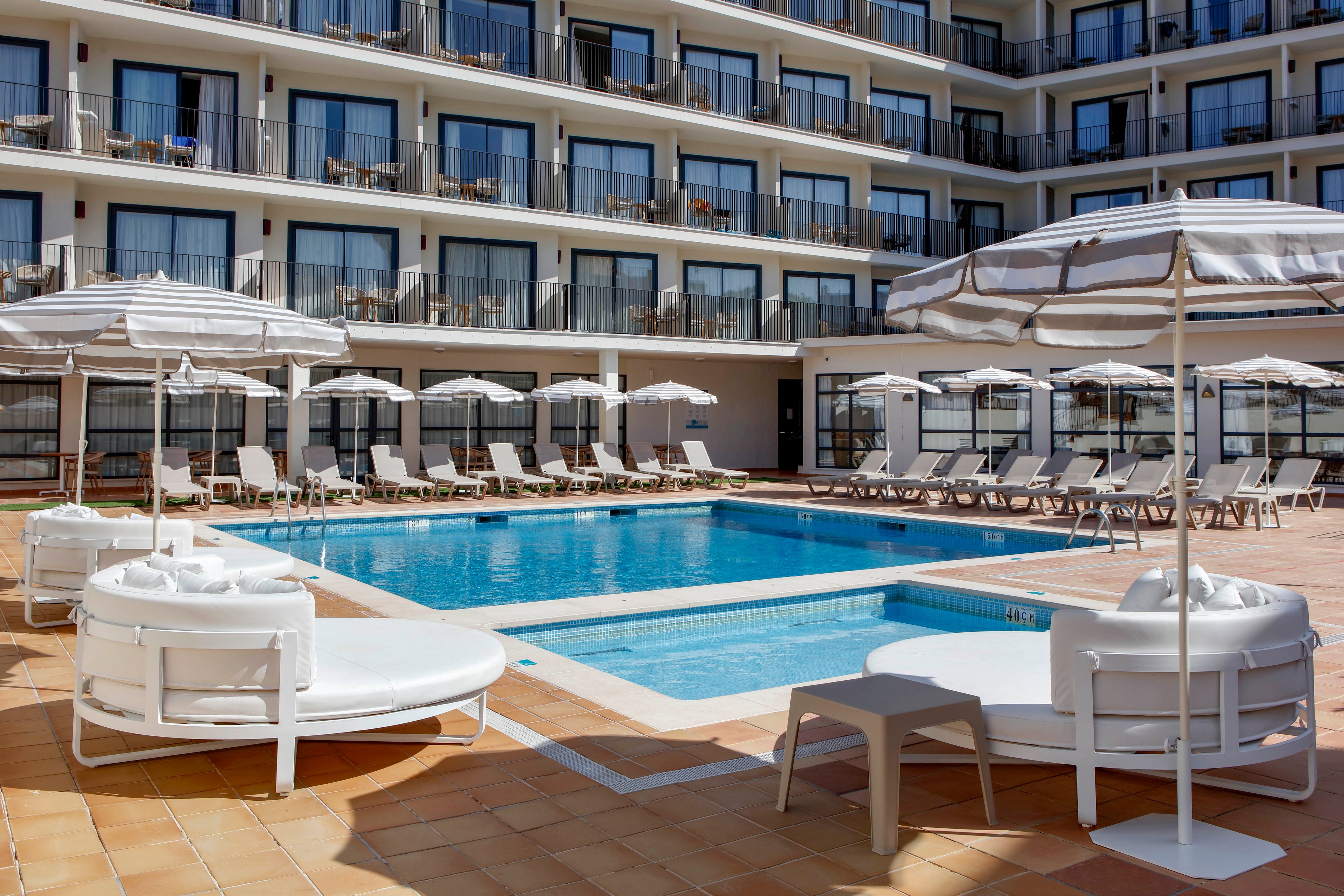 A rectangular outdoor swimming pool sits at the center of a modern hotel courtyard. White sun loungers and round daybeds with striped umbrellas surround the pool on a tiled terrace. The multi-story building in the background features rows of balconies with glass railings and sliding doors. The scene is bright and clean, suggesting a relaxing vacation or resort environment.