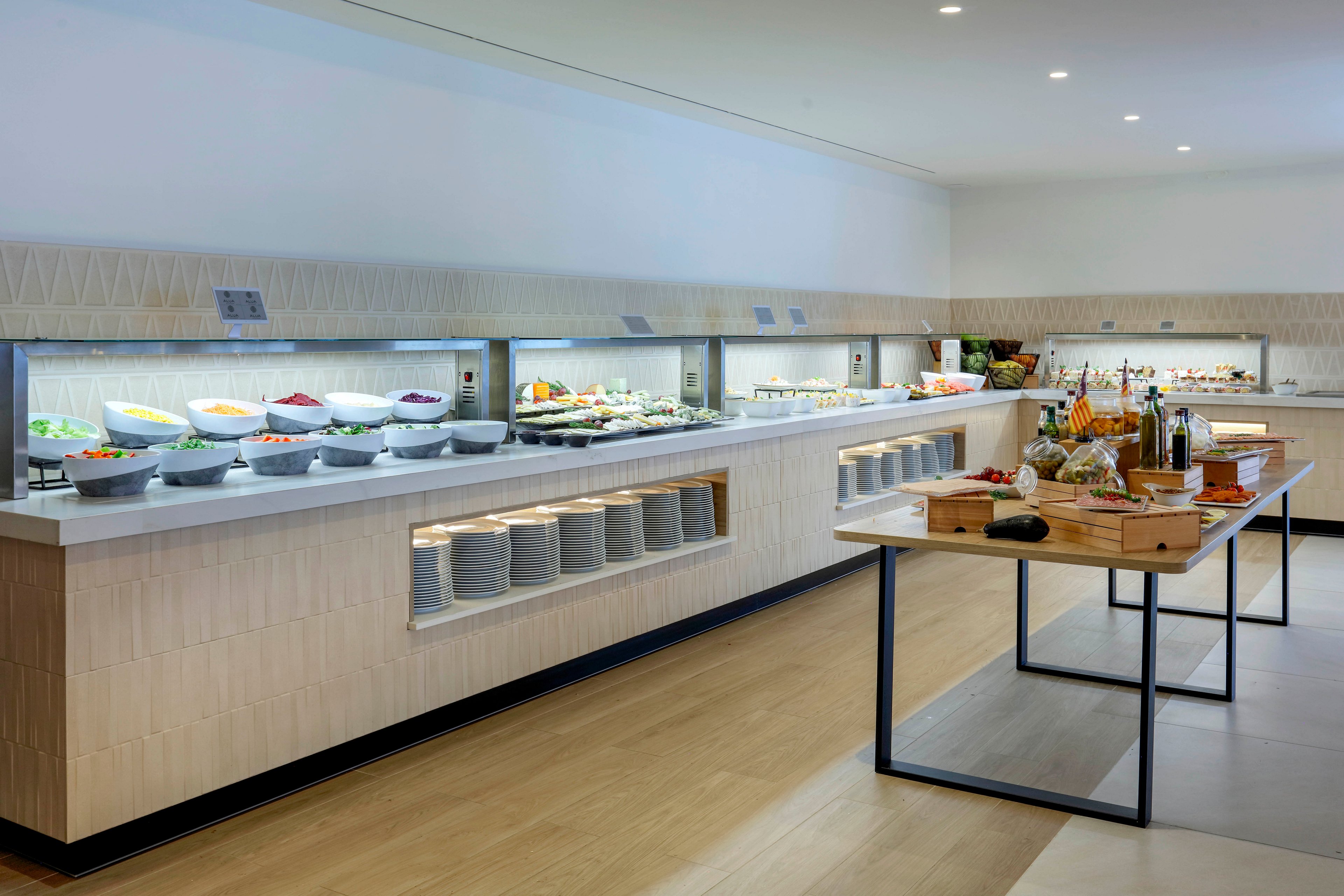 A contemporary self-service buffet counter showcases a wide variety of fresh dishes in white bowls and trays. The setting appears to be a hotel or restaurant dining area with clean, minimalist design and light wood flooring. A separate wooden table in the foreground holds breads, fruits, and condiments arranged in an inviting presentation. Bright, even lighting and neutral tones create a spacious, modern atmosphere focused on food variety and freshness.