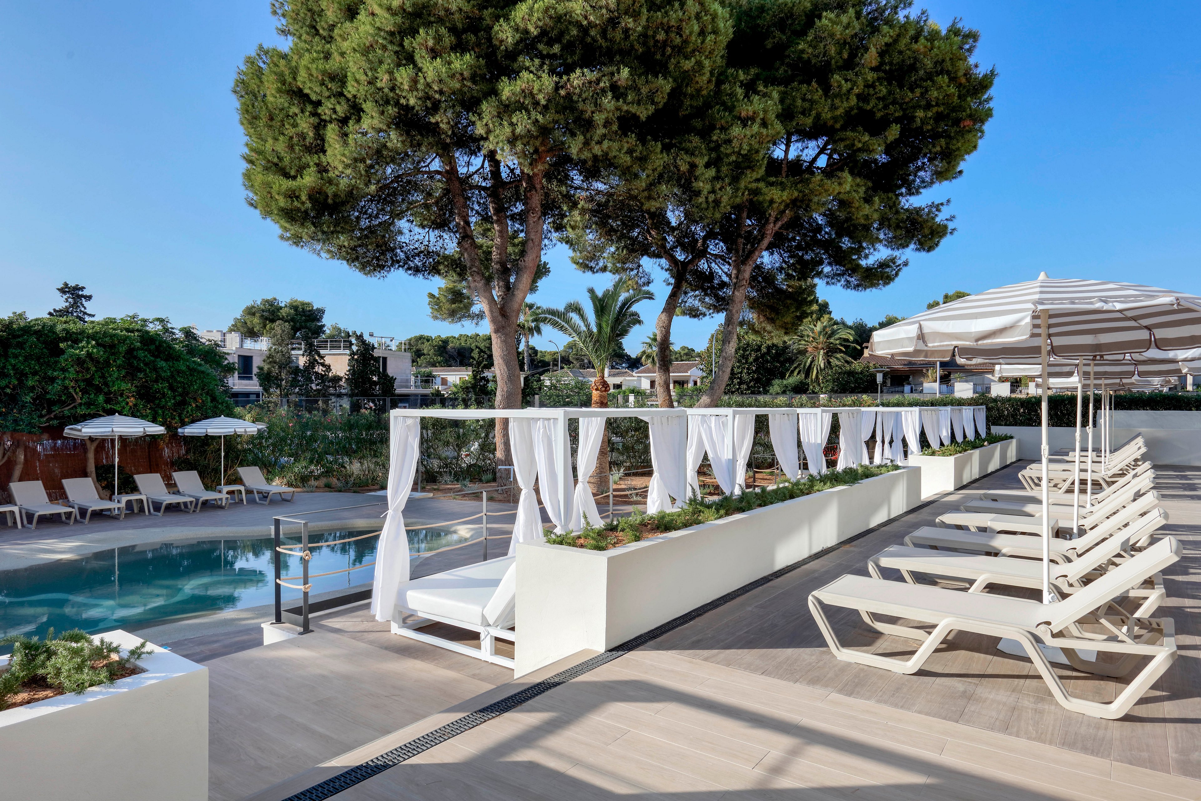 Contemporary poolside area featuring white sun loungers and elegant cabanas with flowing curtains. The setting is an outdoor leisure space surrounded by lush greenery and tall trees under a clear blue sky. A calm swimming pool runs alongside the seating area, with umbrellas providing shade and a relaxed resort atmosphere. The overall scene conveys a clean, minimalist design ideal for vacation, relaxation, and summer recreation.