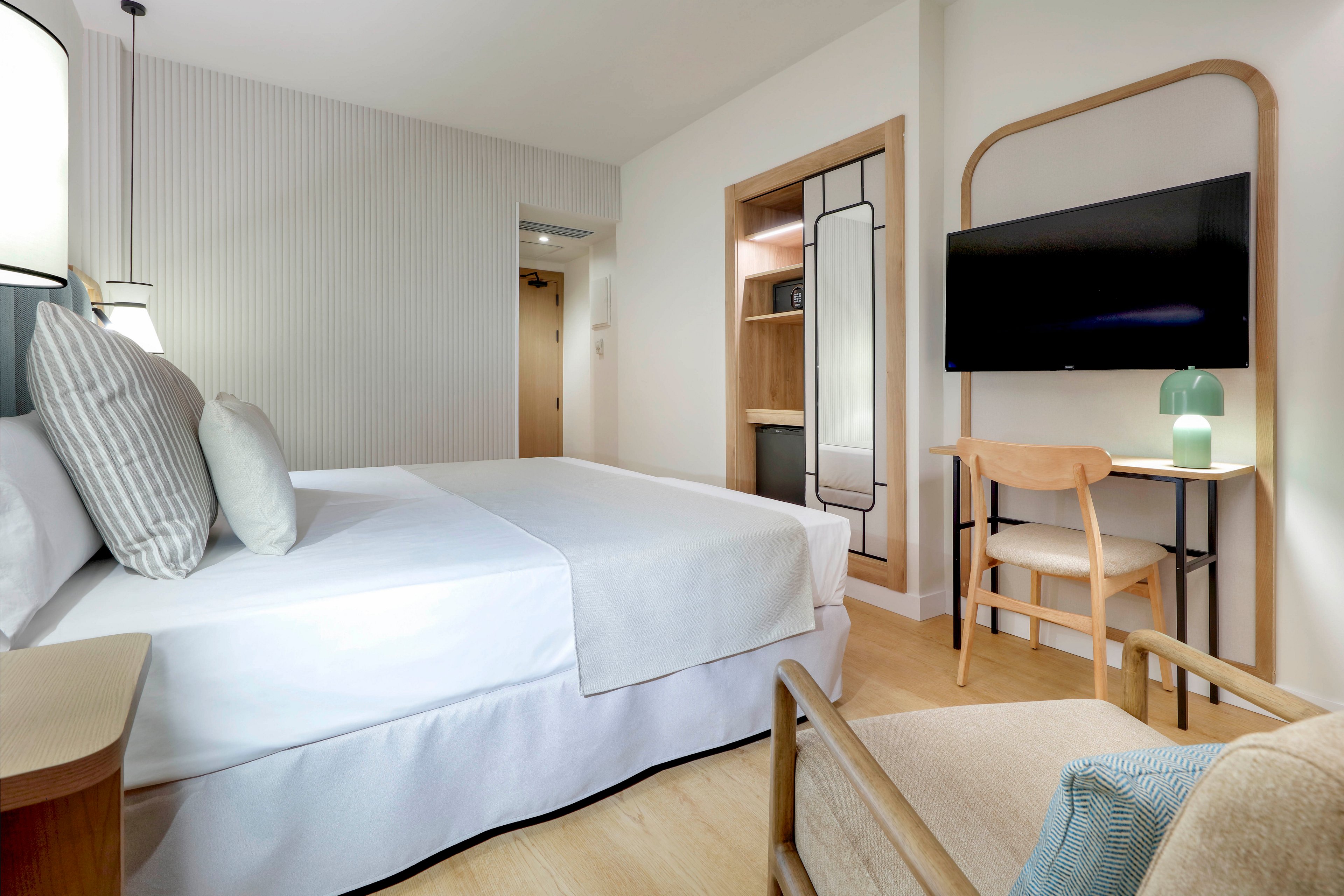 Contemporary hotel-style bedroom featuring a neatly made double bed with white linens and striped pillows. The room includes a wall-mounted flat-screen television above a wooden chair and desk area. Light wood flooring, neutral walls, and soft lighting create a calm, minimalist atmosphere. A small lamp on the desk and an upholstered armchair add comfort and functionality to the space.