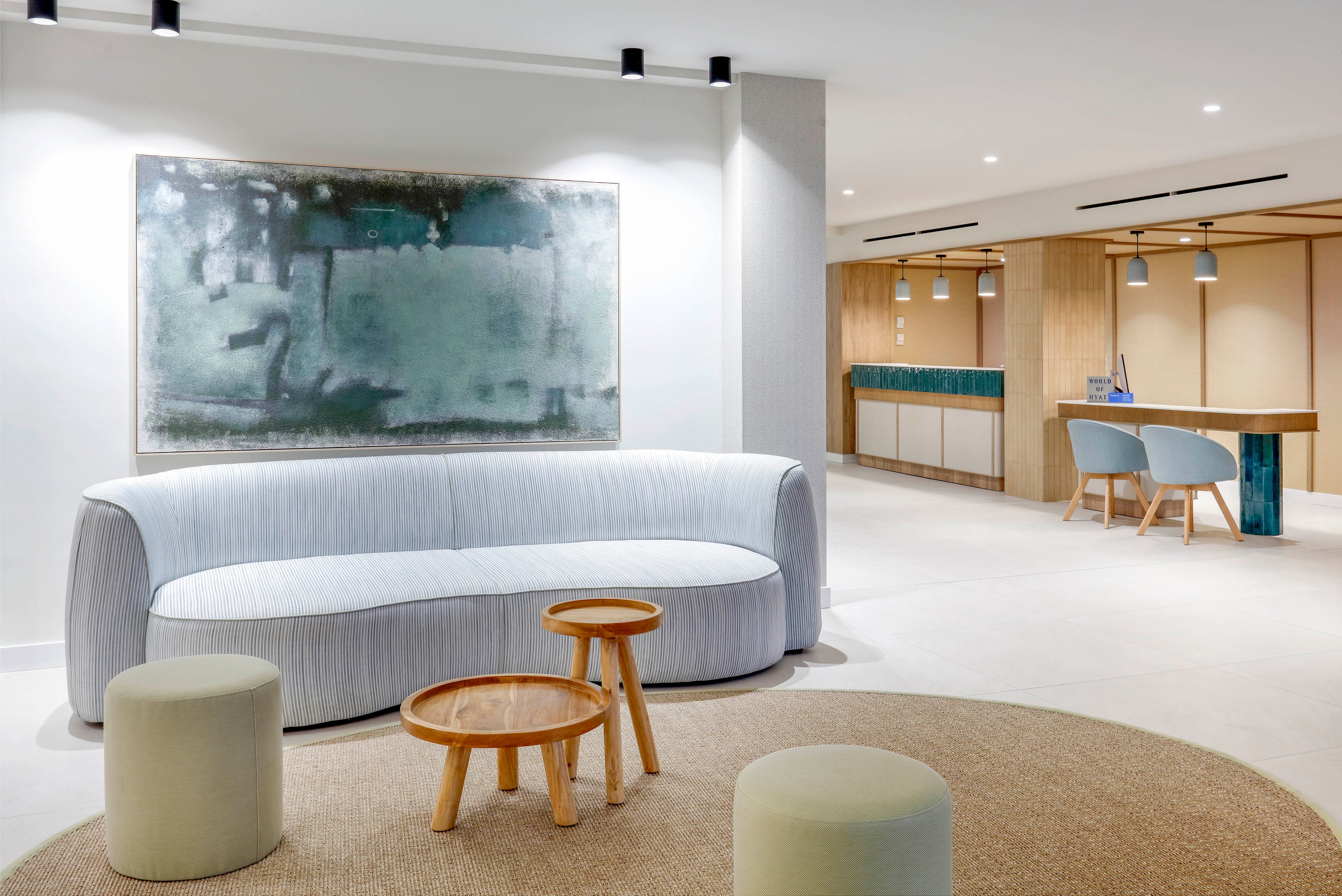 A contemporary lobby features a light gray curved sofa as the central seating element. Two round wooden coffee tables and upholstered stools sit on a large circular rug in the foreground. A large abstract painting hangs on the wall above the sofa, adding subtle color and texture. In the background, a reception or work area with pendant lights, wooden surfaces, and blue chairs completes the clean, minimalist interior design.