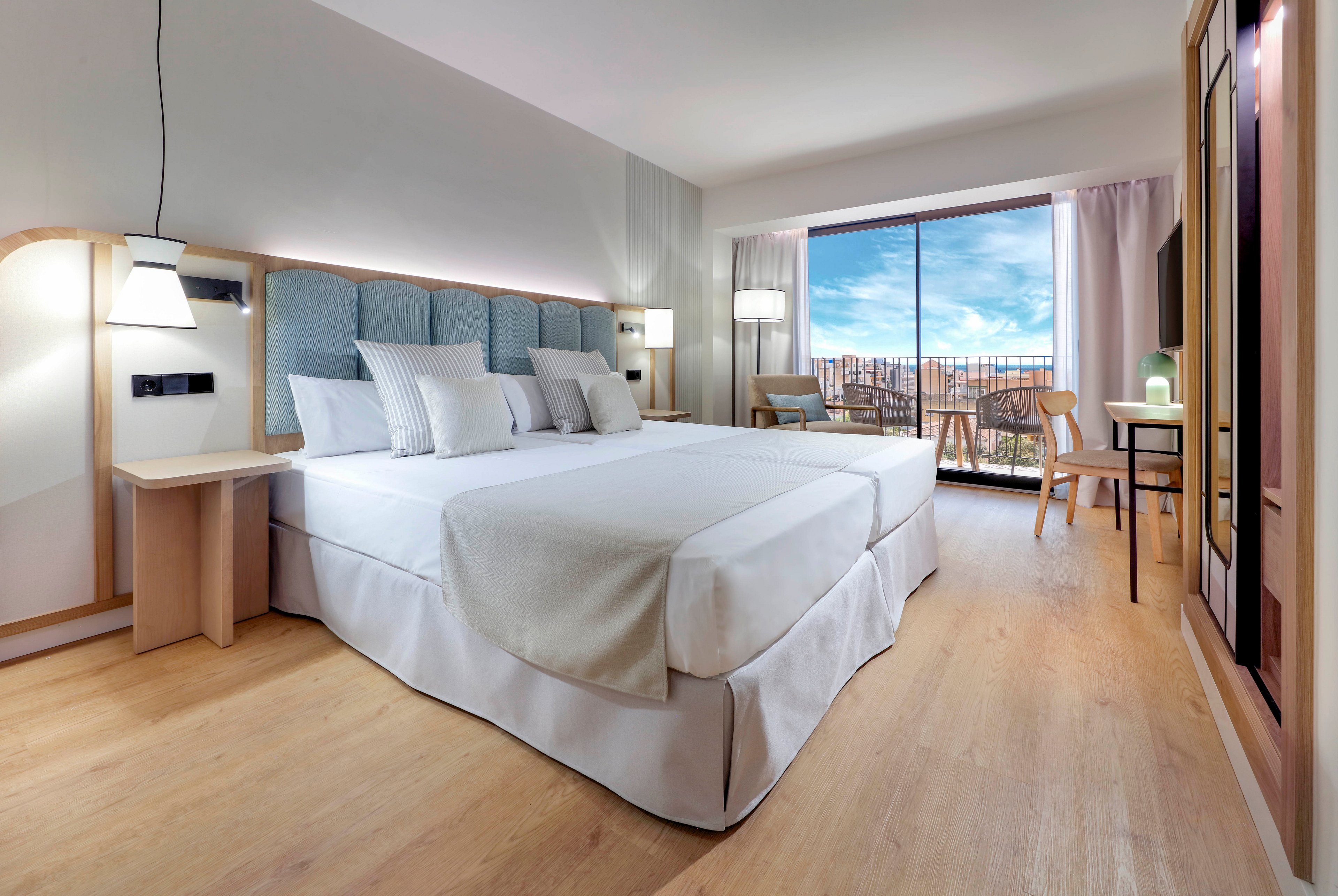 A spacious, contemporary hotel room features a large double bed with white linens and multiple pillows. Light wood flooring and minimalist furniture create a clean, modern aesthetic. A desk and chair sit near a floor-to-ceiling glass door that opens to a balcony with outdoor seating. Natural light floods the room, highlighting the neutral color palette and the blue sky visible outside.
