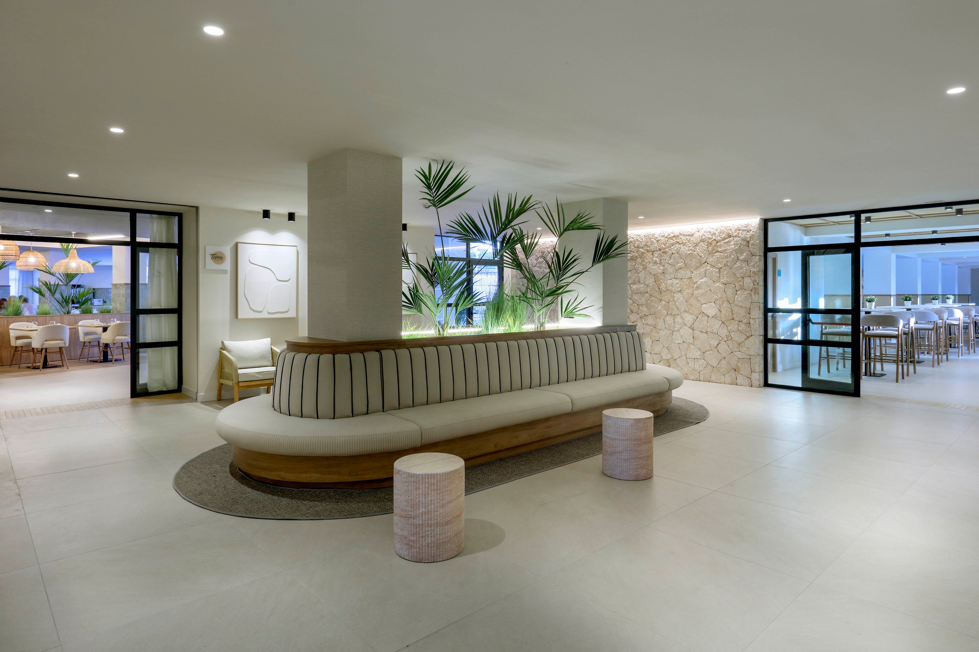 A contemporary hotel lobby features a long curved bench with striped upholstery as the central focal point. The space is bright and minimal, with light stone walls, smooth flooring, and recessed ceiling lights. Potted tropical plants are set behind the bench, adding greenery and contrast to the neutral palette. Glass doors on both sides open to dining areas with wooden chairs and warm pendant lighting, creating an inviting atmosphere.