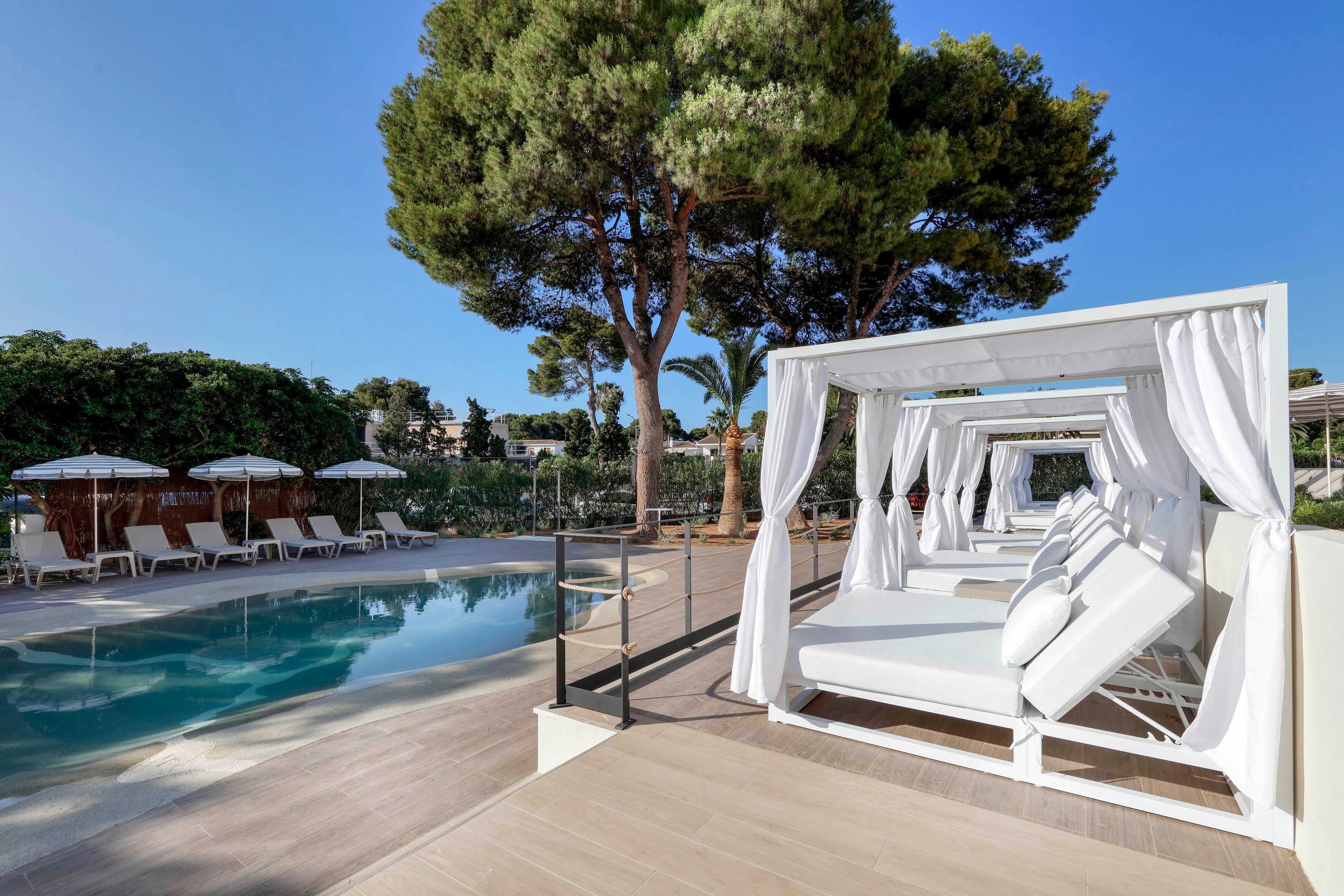 The image shows a row of white canopy daybeds beside a clear outdoor swimming pool. Sun loungers with umbrellas line the opposite side of the pool, framed by lush green trees and shrubs. The scene is brightly lit under a clear blue sky, creating a serene and upscale vacation atmosphere. Clean lines, neutral tones, and natural greenery emphasize relaxation and modern resort style.