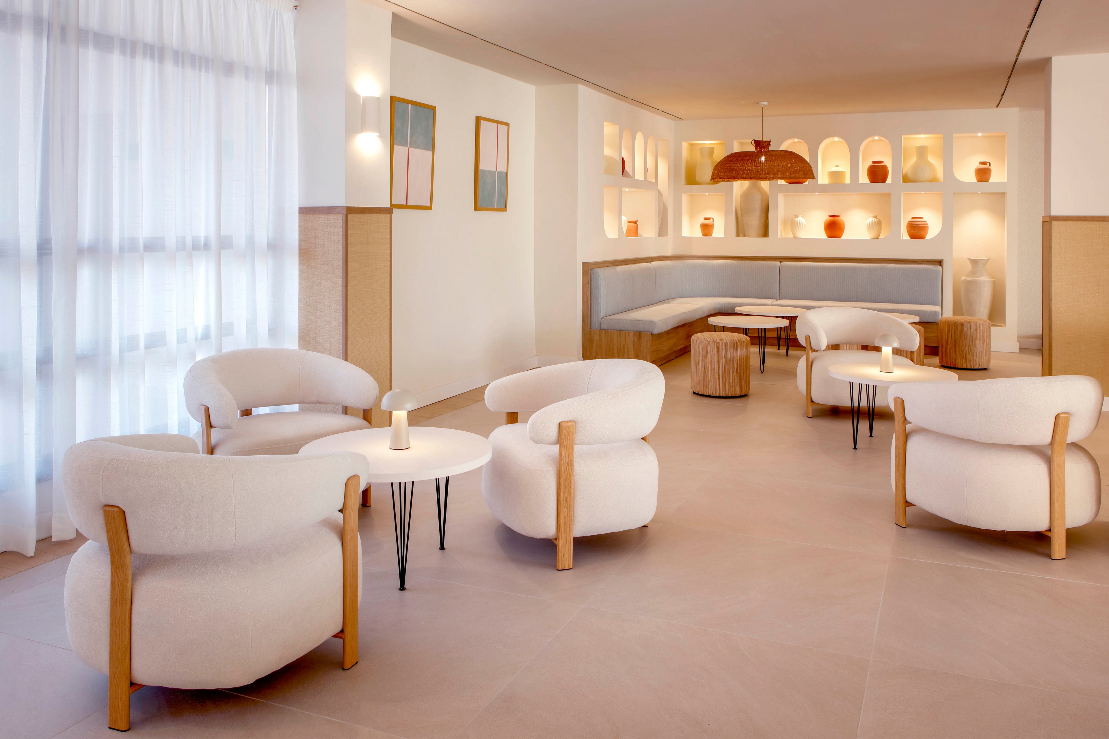 Contemporary lounge area featuring rounded white armchairs arranged around small circular tables. The space is bright and airy, with sheer curtains covering tall windows on one side. Built-in shelving along the back wall displays decorative vases and objects, softly lit to create a warm ambiance. Neutral tones, smooth surfaces, and clean lines emphasize a minimalist, modern interior design.
