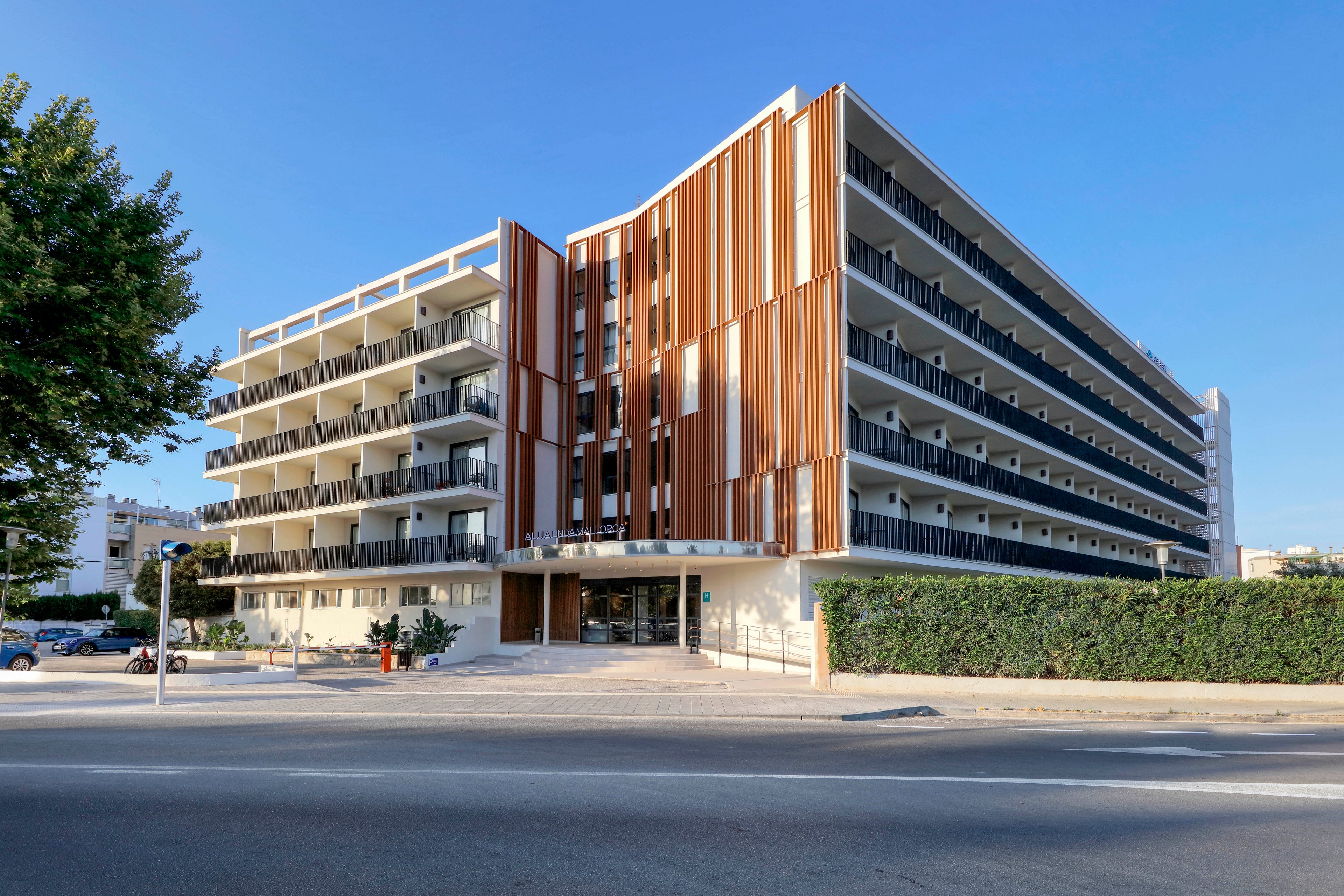 A contemporary multi-story apartment building stands along a wide urban street under a clear blue sky. The structure features long horizontal balconies, vertical wooden slats, and glass railings, giving it a modern architectural appearance. Green hedges and trees frame the property, while the bright daylight emphasizes clean lines and neutral colors. The scene suggests a quiet residential neighborhood with minimal traffic and a calm atmosphere.