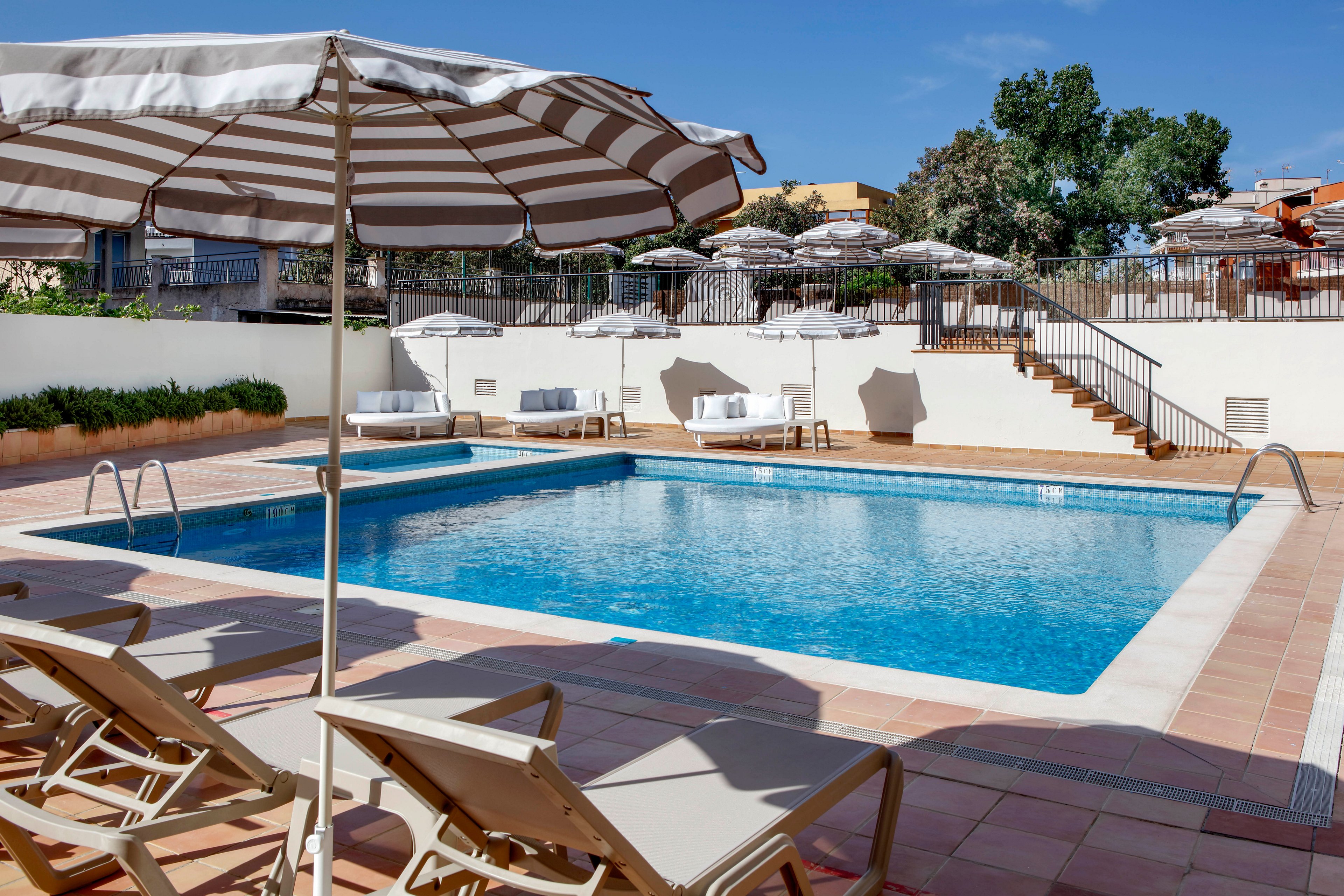 An outdoor rectangular swimming pool is surrounded by sun loungers and large striped umbrellas under a clear blue sky. The setting appears to be a hotel or resort terrace with tiled flooring and white walls. Stairs and railings lead up to an elevated area with additional seating and umbrellas. The scene is bright and inviting, suggesting a relaxing vacation atmosphere.