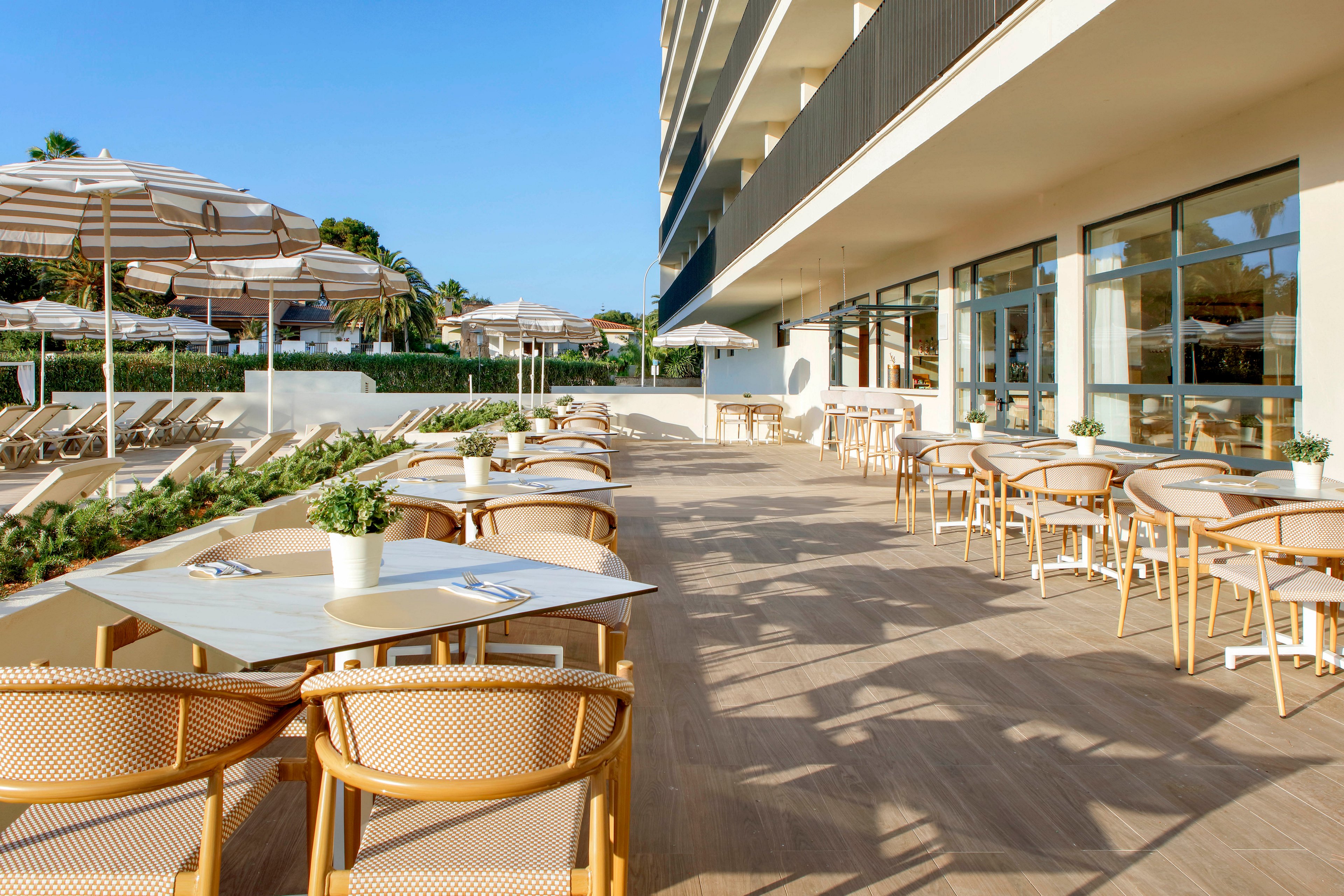 A spacious hotel terrace features neatly arranged outdoor dining tables and chairs in warm natural tones. The setting overlooks a poolside area with sun loungers and striped umbrellas under a clear blue sky. Large windows line the building, reflecting the bright daylight and enhancing the open, airy atmosphere. Potted plants on the tables add a touch of greenery and complement the relaxed vacation vibe.