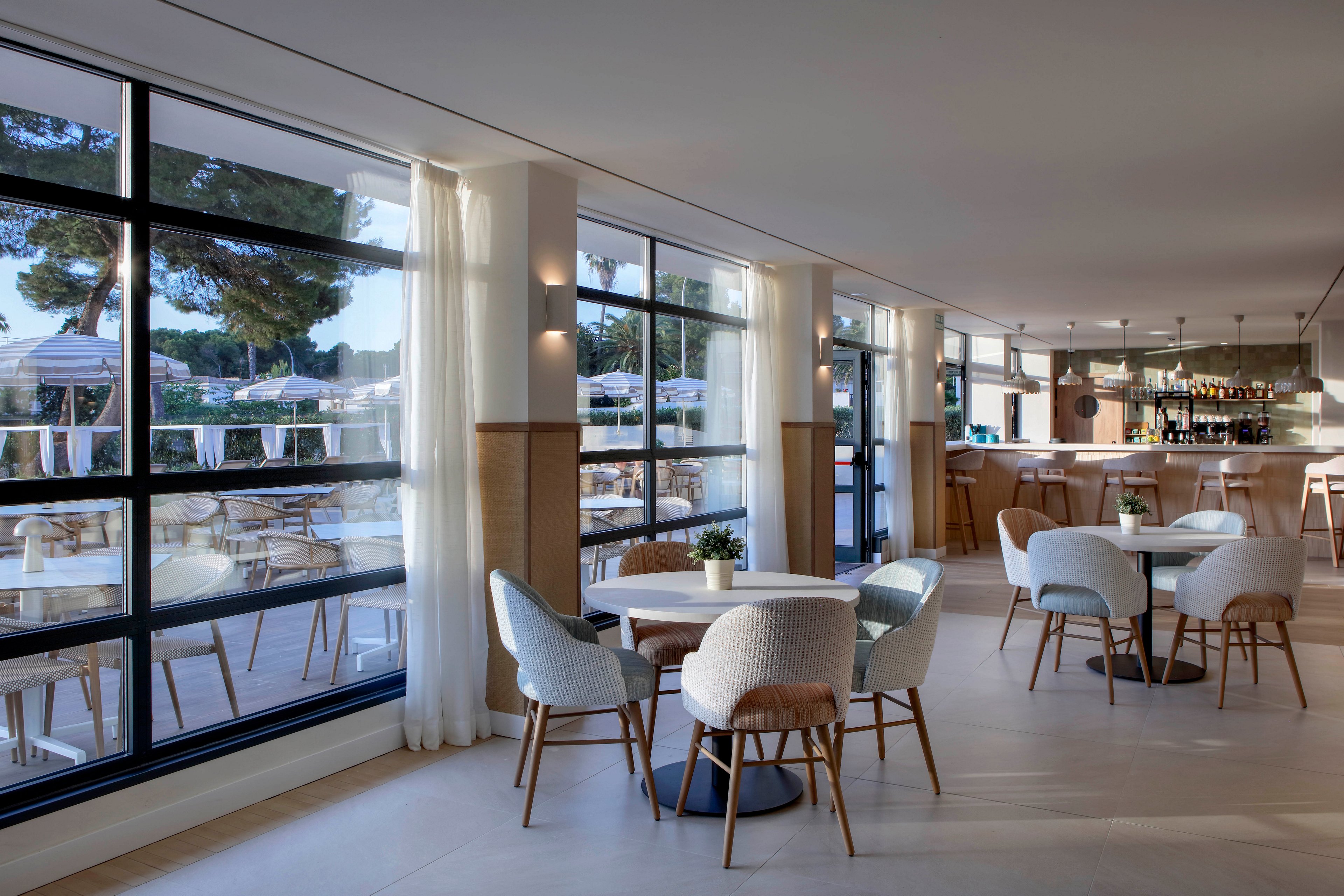 Bright contemporary restaurant interior features light wood floors and neutral upholstered chairs around small tables. Floor-to-ceiling windows reveal an outdoor terrace with additional seating and views of trees and the sea. Soft natural light fills the space, complemented by wall sconces and a sleek bar area in the background. The overall atmosphere is calm, airy, and inviting for casual dining or drinks.