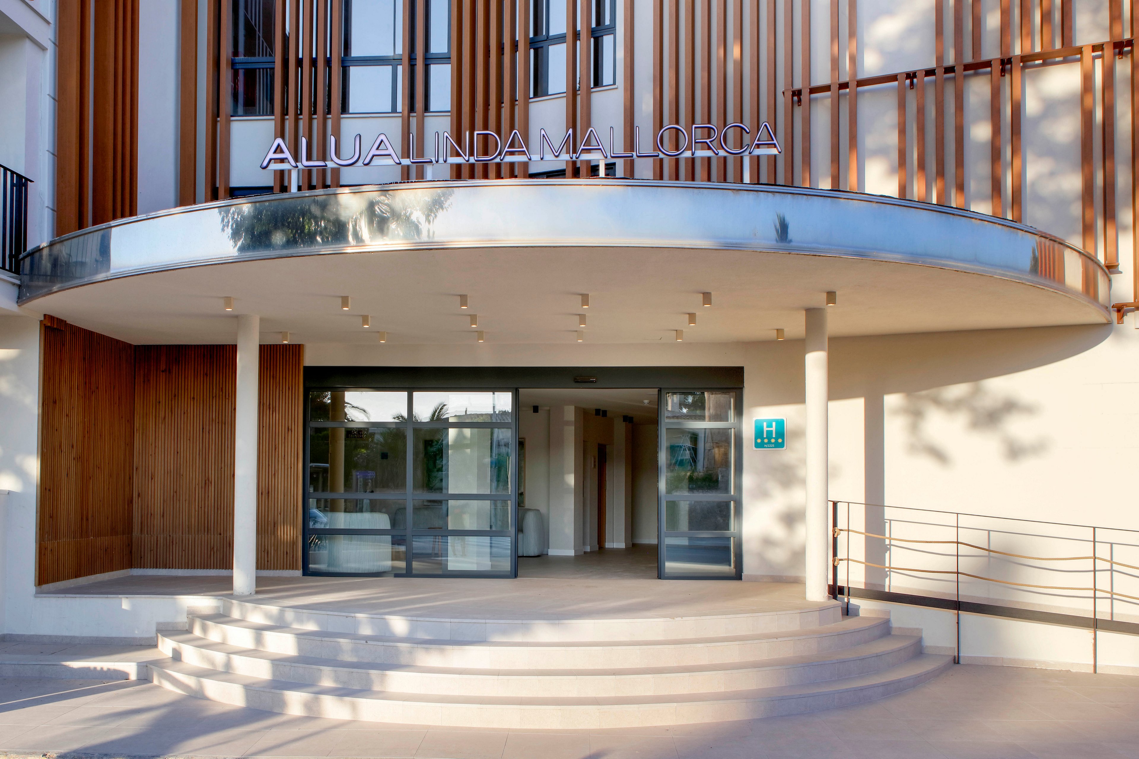 The image shows the main entrance of the Alua Linda Mallorca hotel with a curved canopy and wide steps leading up to glass doors. Vertical wooden slats decorate the facade, adding a contemporary architectural touch. A blue hotel rating plaque is visible beside the entrance, and a metal handrail lines the ramp on the right. Soft daylight illuminates the clean, minimalist exterior, creating a welcoming atmosphere.
