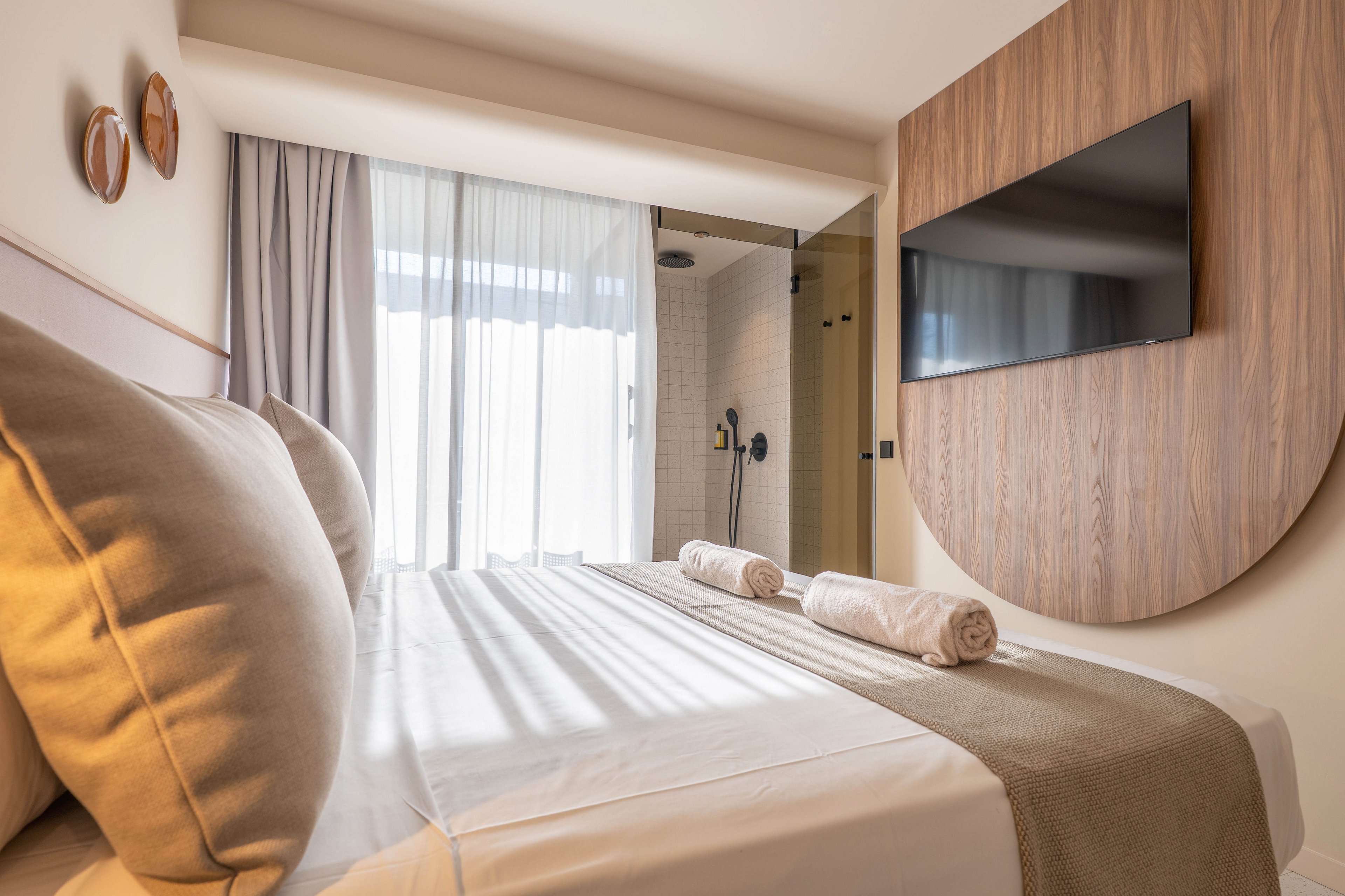 A contemporary hotel room features a neatly made bed with two rolled towels placed on top. The room is decorated in neutral tones with a large flat-screen TV mounted on a wooden accent wall. Natural light streams in through sheer curtains, and a glass-walled shower is visible in the background, enhancing the open, airy feel.