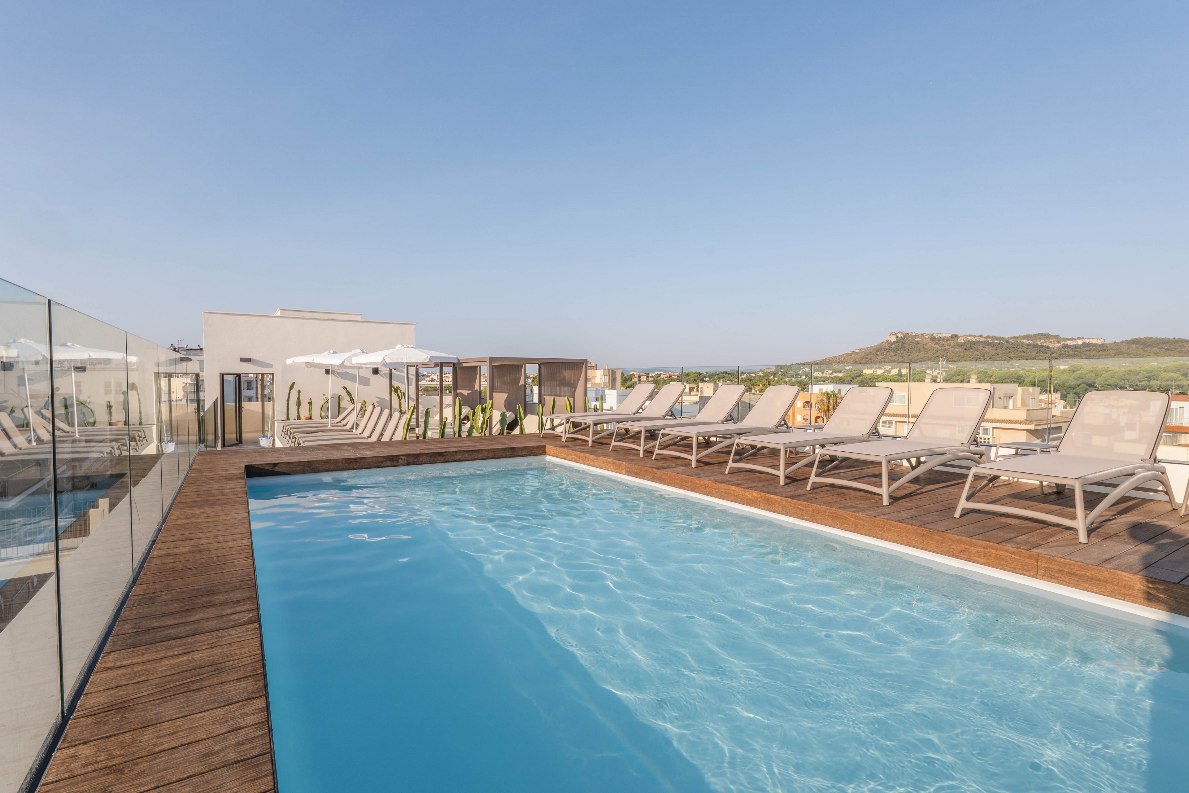 A contemporary rooftop pool is surrounded by wooden decking and lined with multiple sun loungers and white umbrellas. The setting is bright and open, with clear blue water reflecting the sunny sky. Glass railings provide safety while offering unobstructed views of distant hills and urban landscape. The overall mood is relaxed and inviting, ideal for leisure and sunbathing.