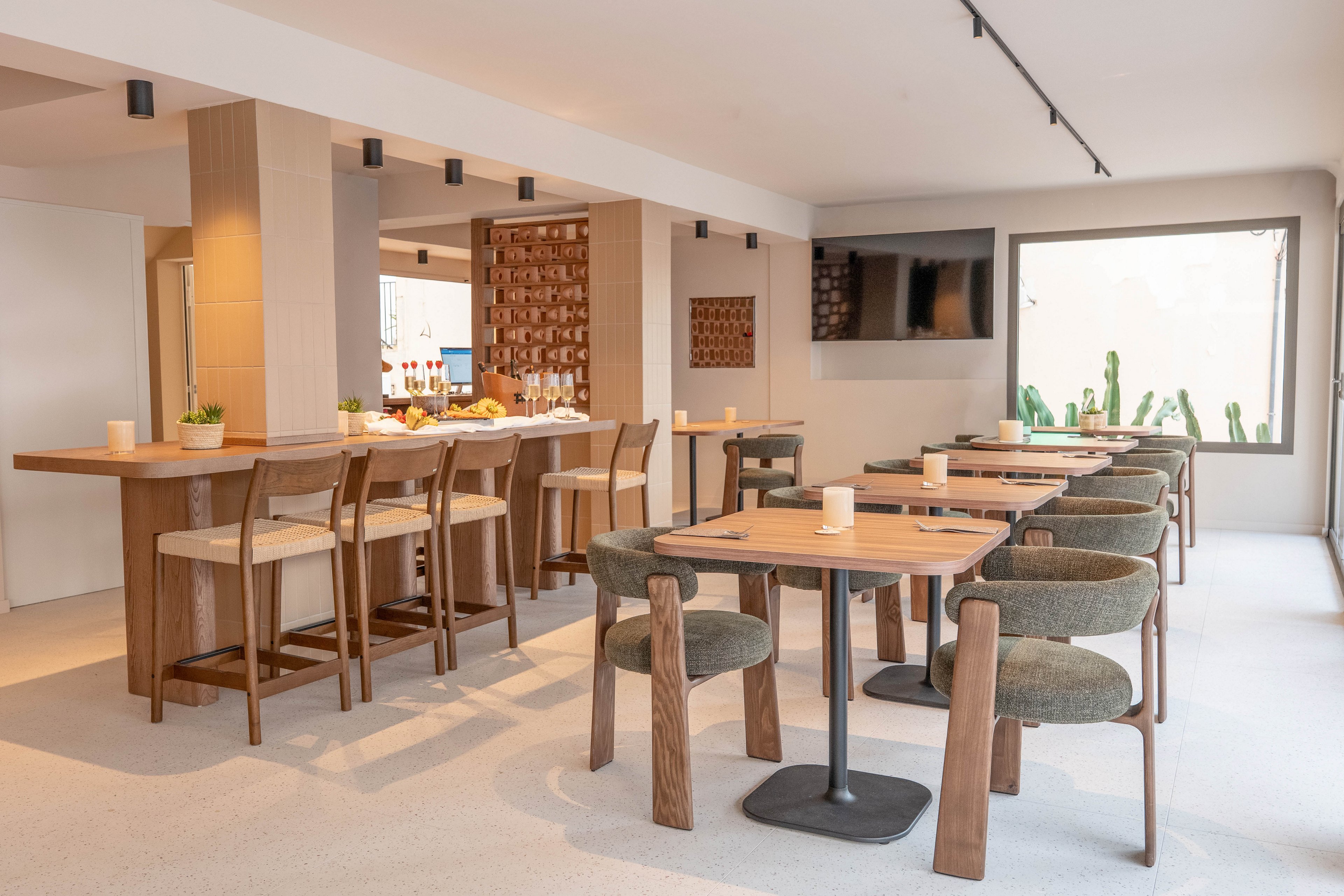 A contemporary restaurant dining area features wooden tables and chairs arranged neatly in a bright, minimalist space. The setting includes a bar counter with stools, a large window letting in natural light, and a wall-mounted television. Neutral tones and clean lines create a calm, inviting atmosphere, with small decorative plants adding a touch of greenery. No visible text or numbers are present in the image.