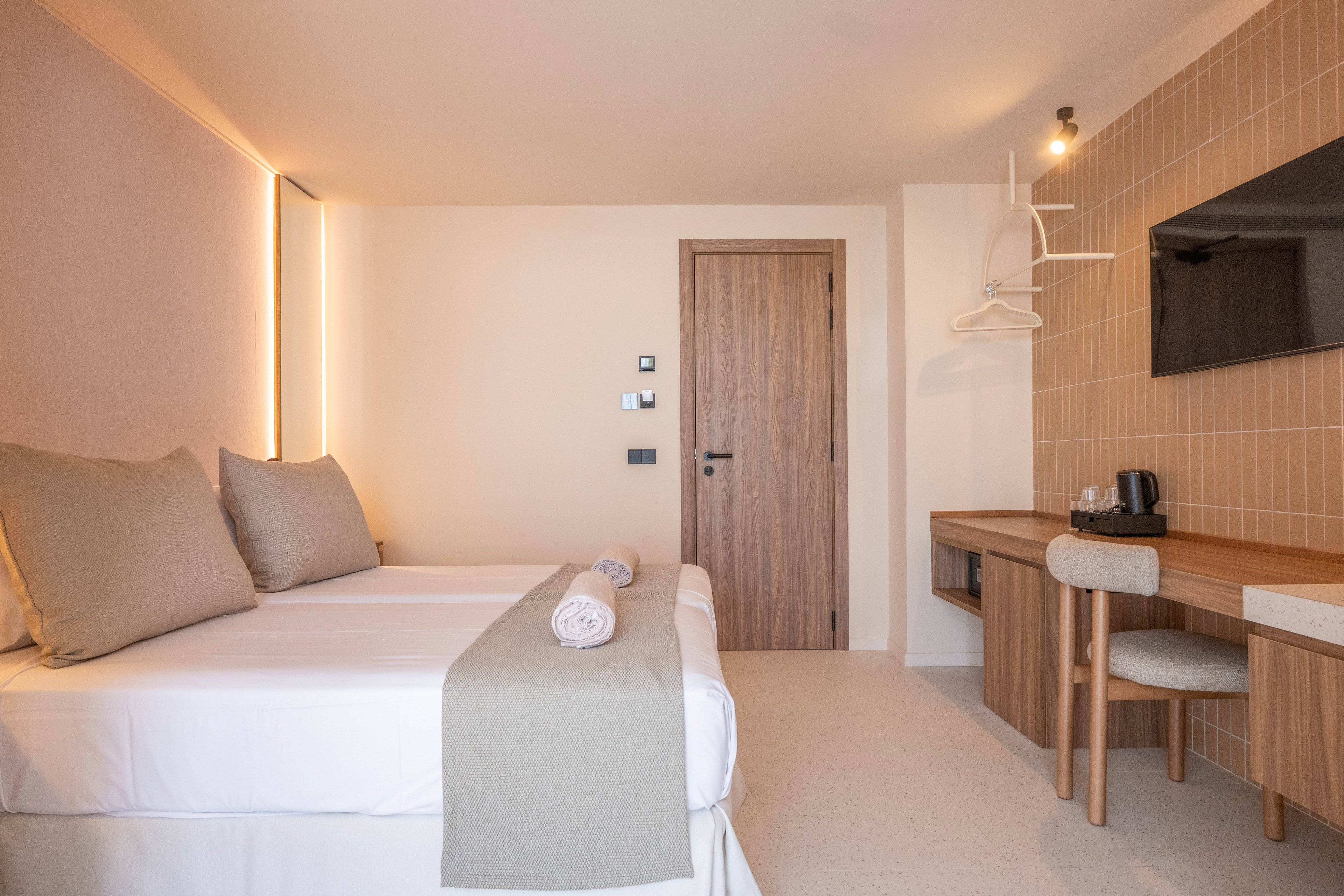 A contemporary hotel room features a double bed with neutral bedding and two rolled towels placed on top. The space includes a wooden desk with a chair, a mounted flat-screen television, and a coffee station. The room is decorated in soft beige and wood tones, with clean lines and minimal decor, creating a calm and inviting atmosphere. No visible text or numeric values are present in the image.