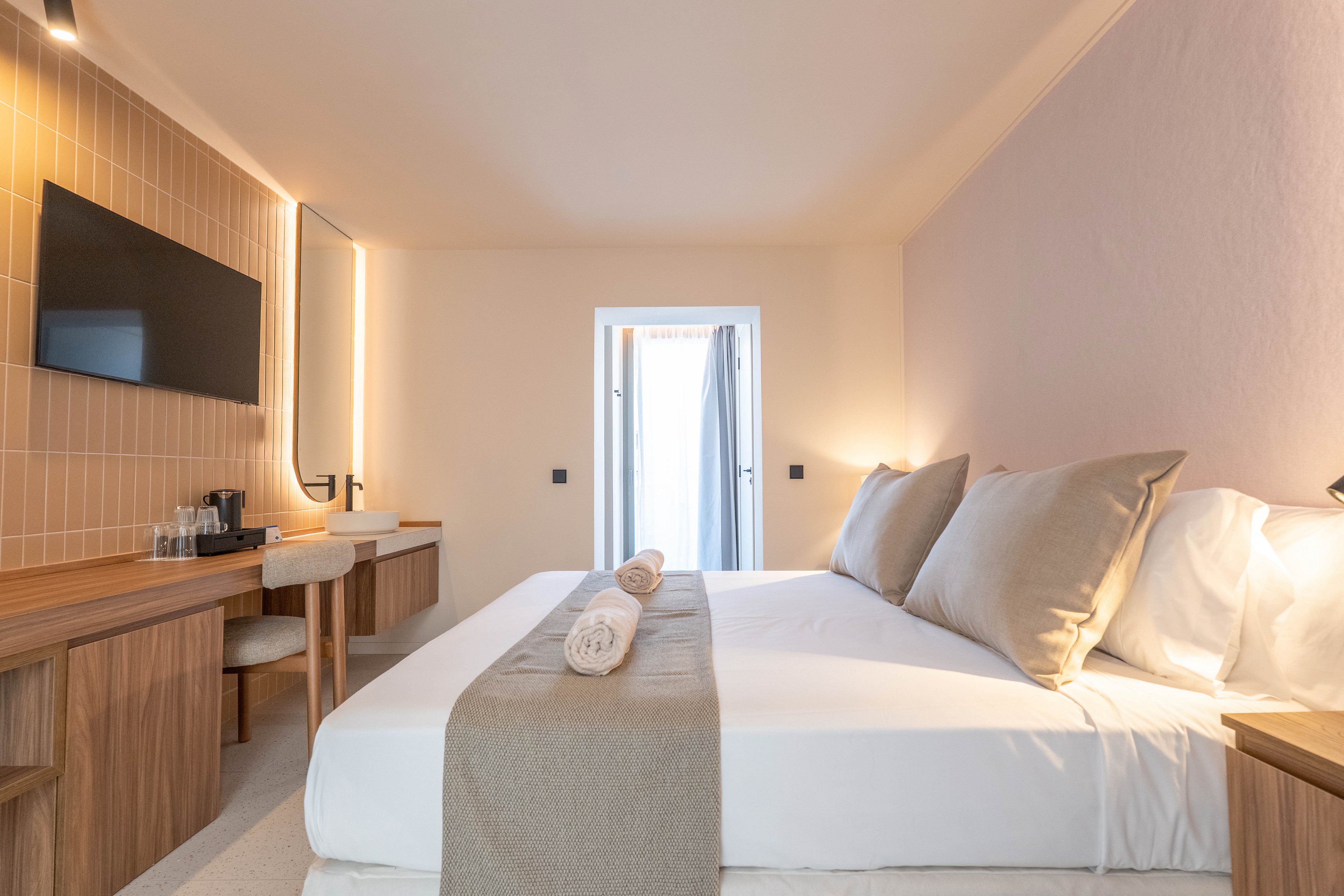 A bright, contemporary hotel room features a large bed with neutral bedding and two rolled towels placed on top. The space includes a wooden desk, chair, flat-screen TV, and a coffee station, all set against soft beige and pastel walls. Natural light enters through a glass door, creating a calm and inviting atmosphere.