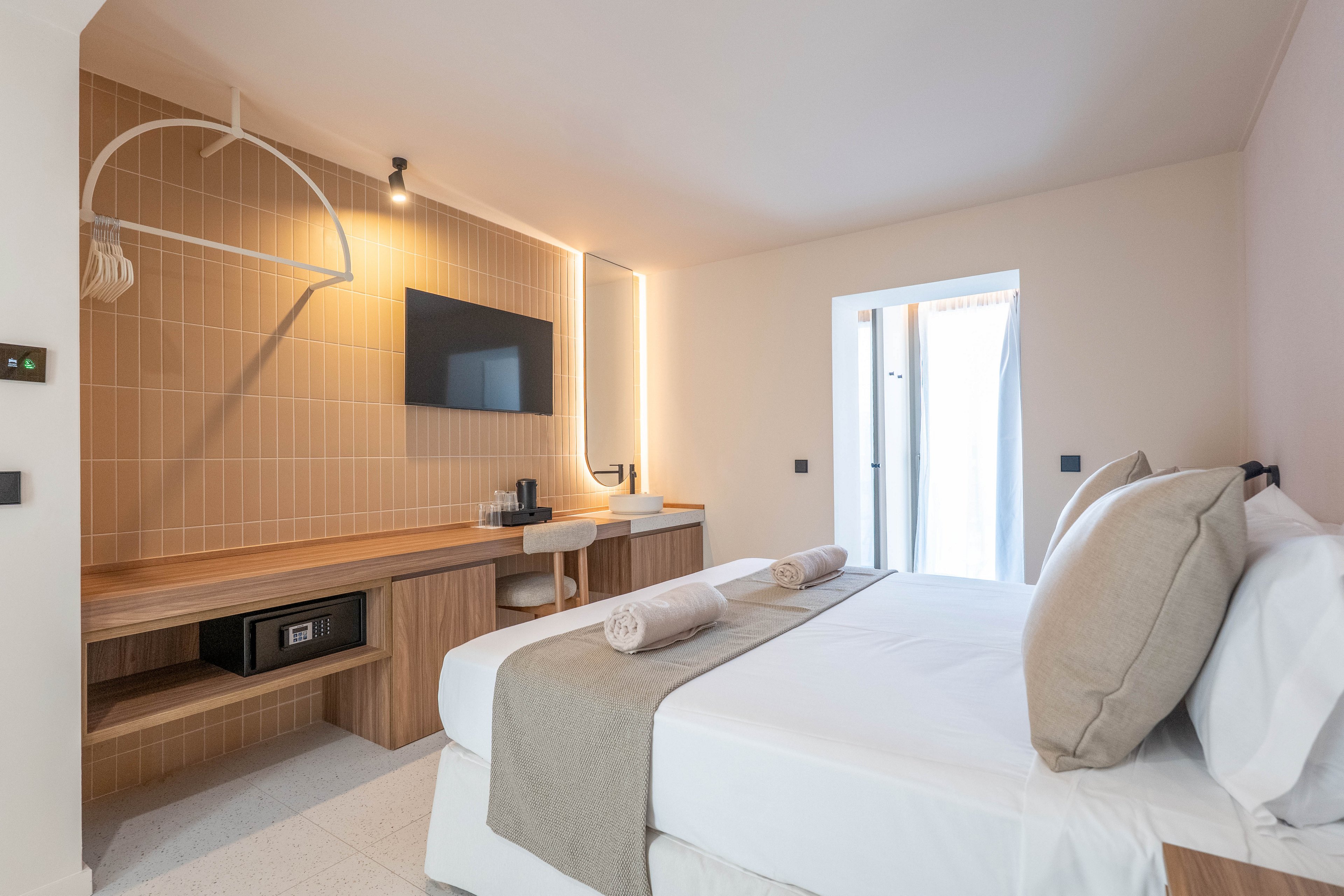 A contemporary hotel room features a neatly made bed with beige and white linens, two rolled towels, and large pillows. The room includes a wooden desk, wall-mounted flat-screen TV, and open closet with a safe. Natural light enters through a small balcony door, highlighting the clean, minimalist design and soft neutral color palette.