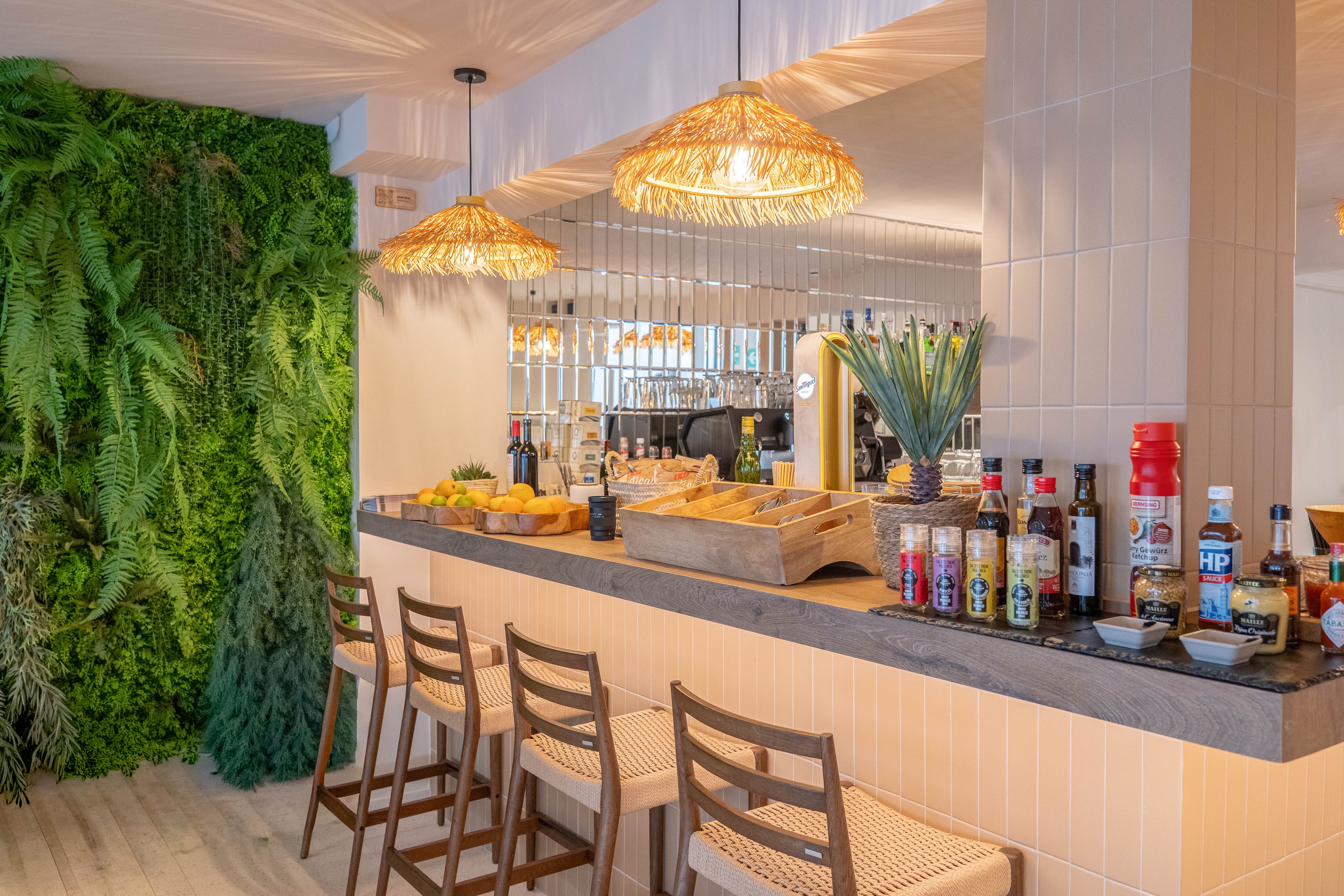 A contemporary bar area features a sleek stone countertop with various condiment bottles and sauces clearly displayed. The setting includes woven bar stools, pendant lights with natural fiber shades, and a lush green plant wall adding a fresh ambiance. Reflections in the mirrored backsplash enhance the spacious feel, while bread baskets and fruit add a casual touch. No visible numeric values or explicit brand names are present.