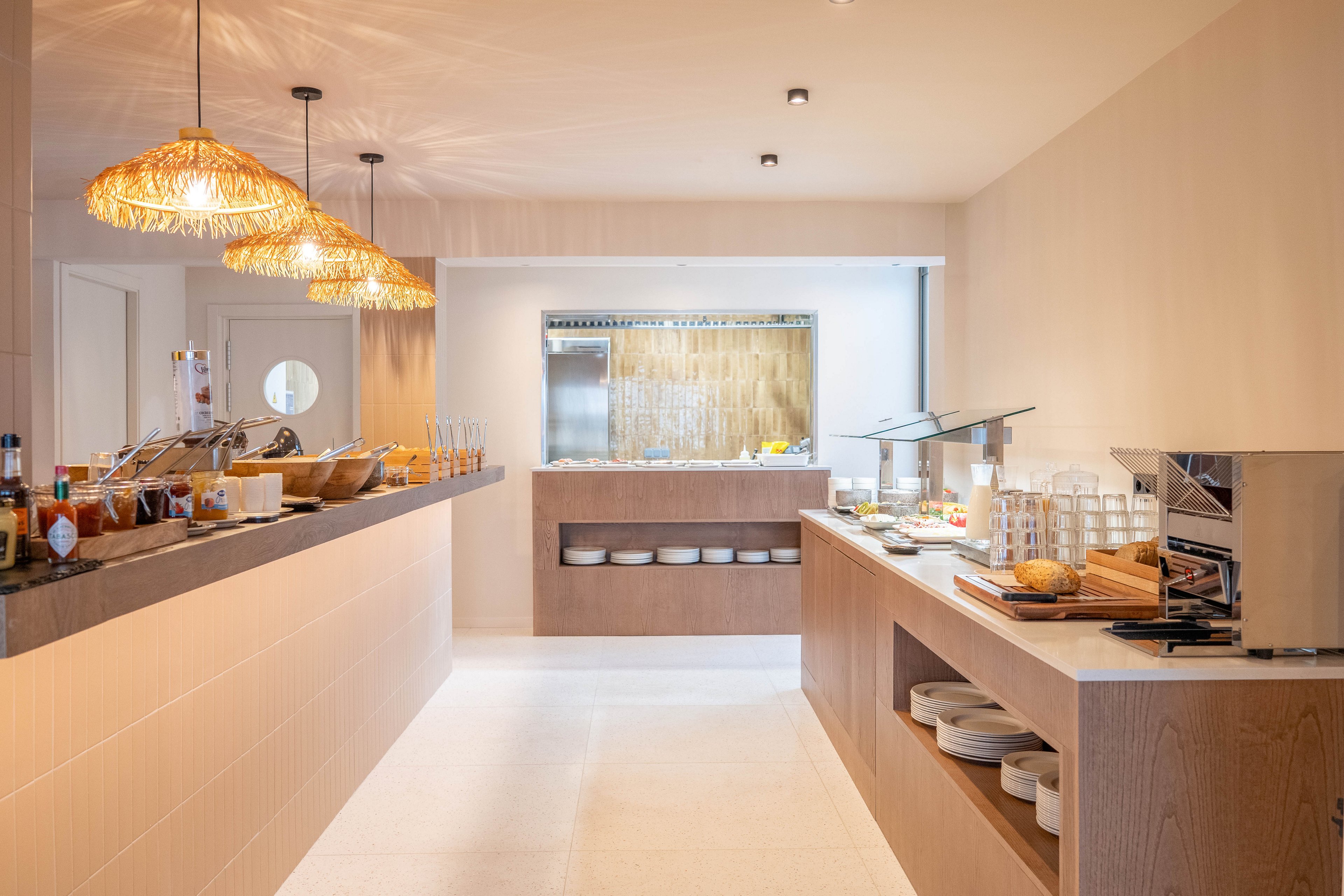A contemporary breakfast buffet is set up in a bright hotel dining area. The scene features wooden counters, open shelving with plates, and a variety of breakfast foods and beverages. Woven pendant lights hang above the counter, adding a warm, inviting atmosphere. The overall palette is neutral with natural textures, and no people are present in the image.