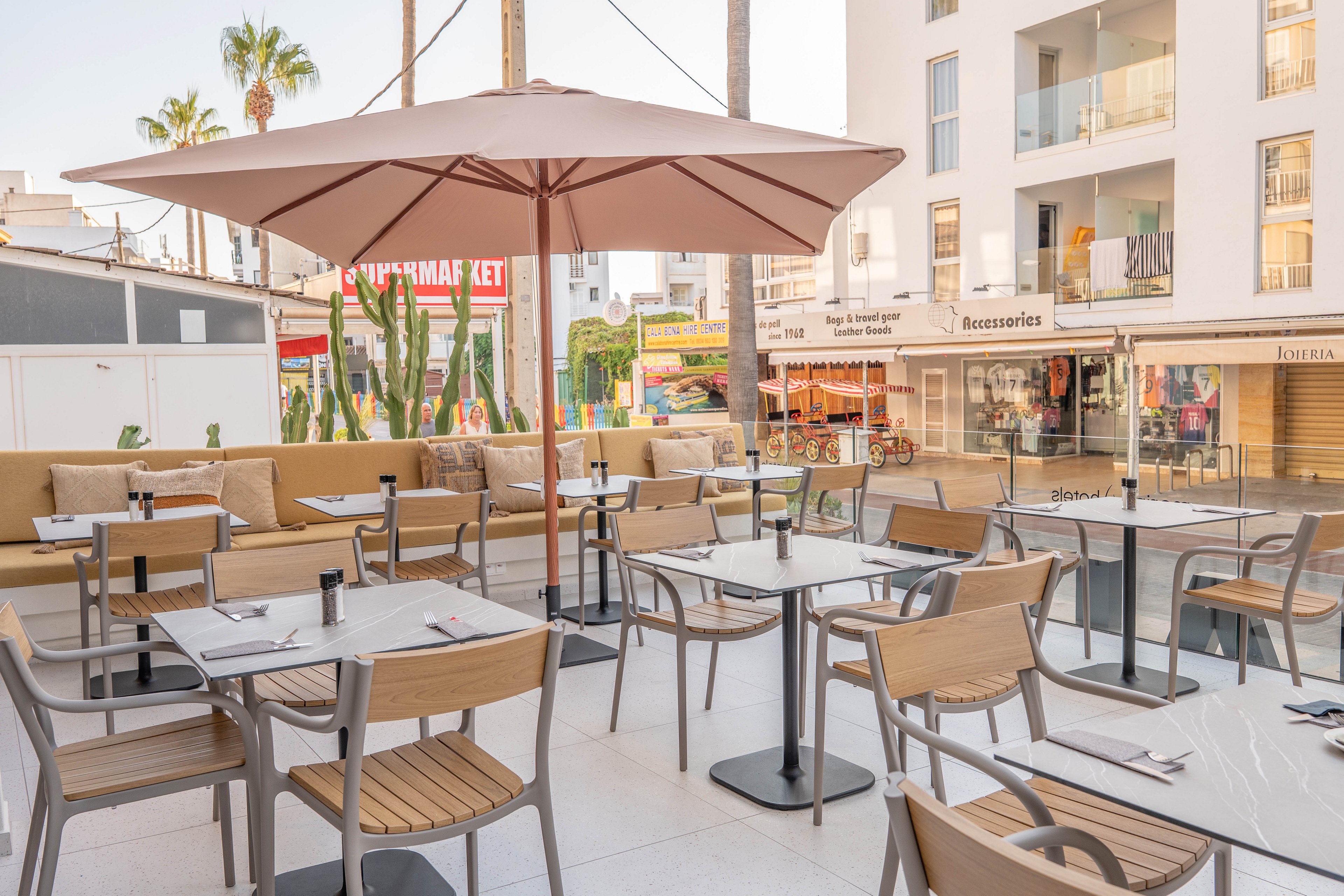A contemporary outdoor café features neatly arranged tables and chairs under a large beige umbrella. The setting is a sunny urban plaza with palm trees, surrounded by modern buildings and visible storefronts, including a market and accessories shop. The atmosphere is relaxed and inviting, with neutral tones and clean lines. No people are present, highlighting the tranquil early morning or off-peak ambiance.