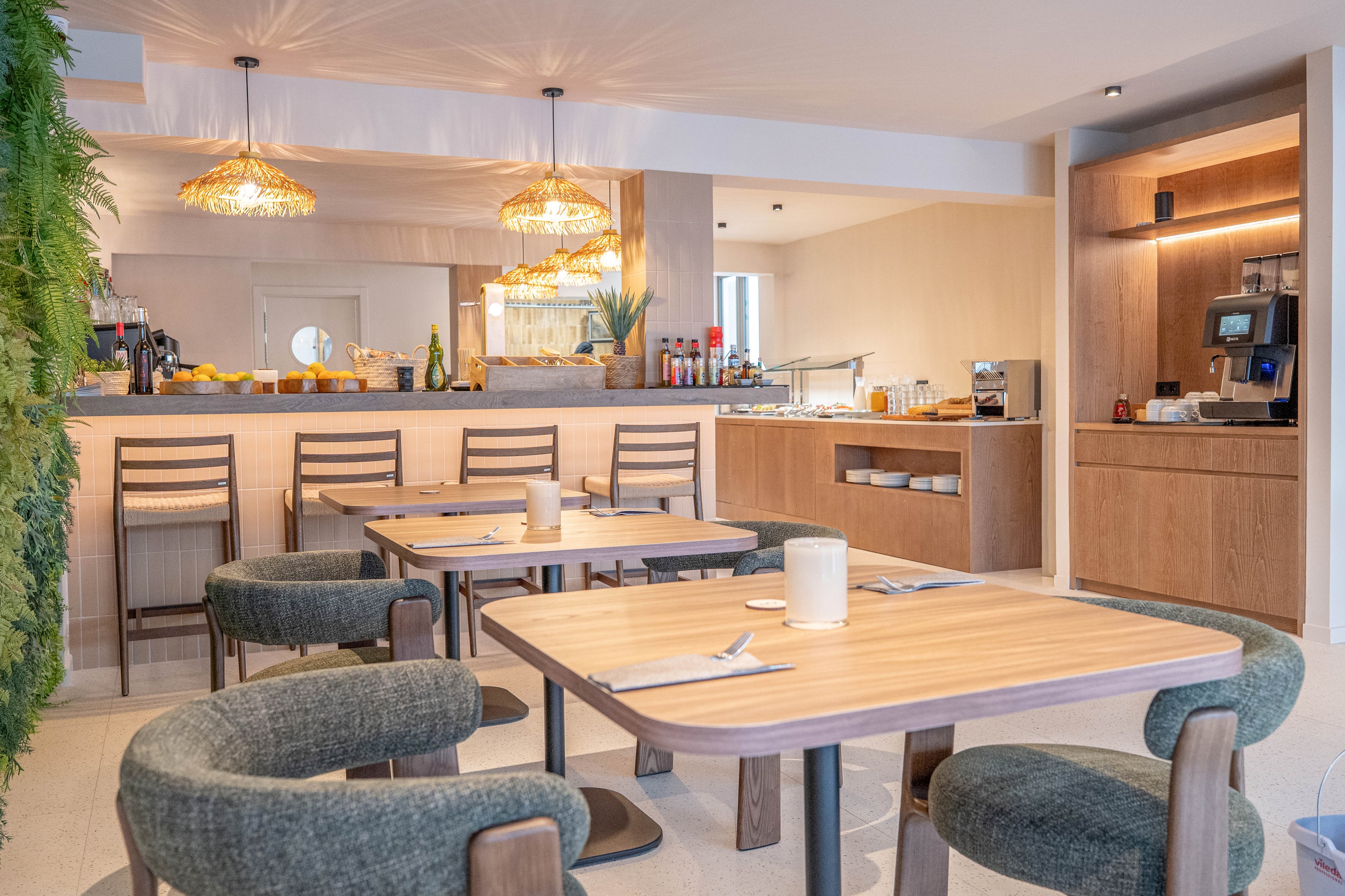 A contemporary dining space features wooden tables and cushioned chairs arranged neatly in a bright, inviting breakfast area. The setting includes a buffet counter with various food items, drinks, and a coffee machine, all under warm pendant lighting. Greenery on the wall and minimalist decor create a relaxed, welcoming atmosphere. No visible people or explicit text are present in the image.
