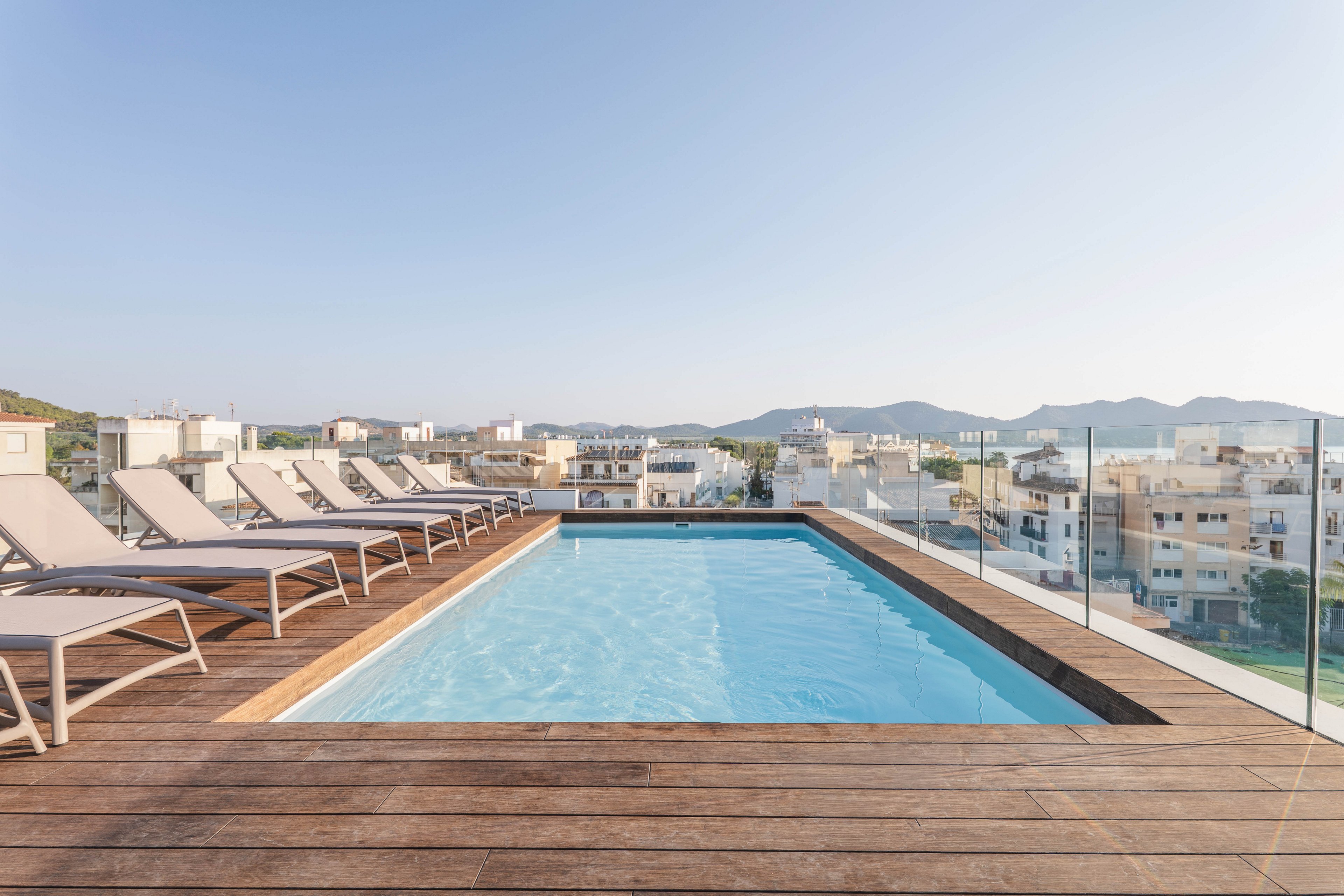 A rectangular rooftop swimming pool is surrounded by wooden decking and several lounge chairs, all set against a backdrop of a cityscape and distant mountains. The pool area is enclosed with clear glass railings, providing an unobstructed view. The scene is bathed in natural daylight, creating a serene and inviting atmosphere. No visible people or text are present in the image.
