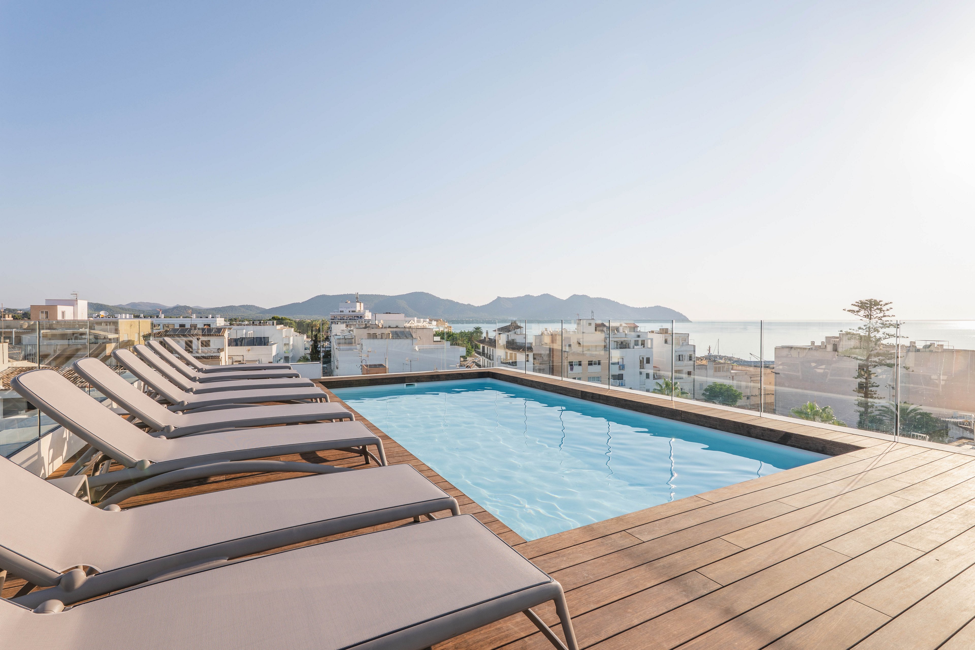 A modern rooftop swimming pool is surrounded by several empty lounge chairs, offering a relaxing spot with panoramic views of a coastal city. The setting is bright and airy, with clear skies and distant mountains visible. The pool area features wooden decking and glass railings, creating a contemporary and inviting atmosphere. No visible text or numbers are present in the image.