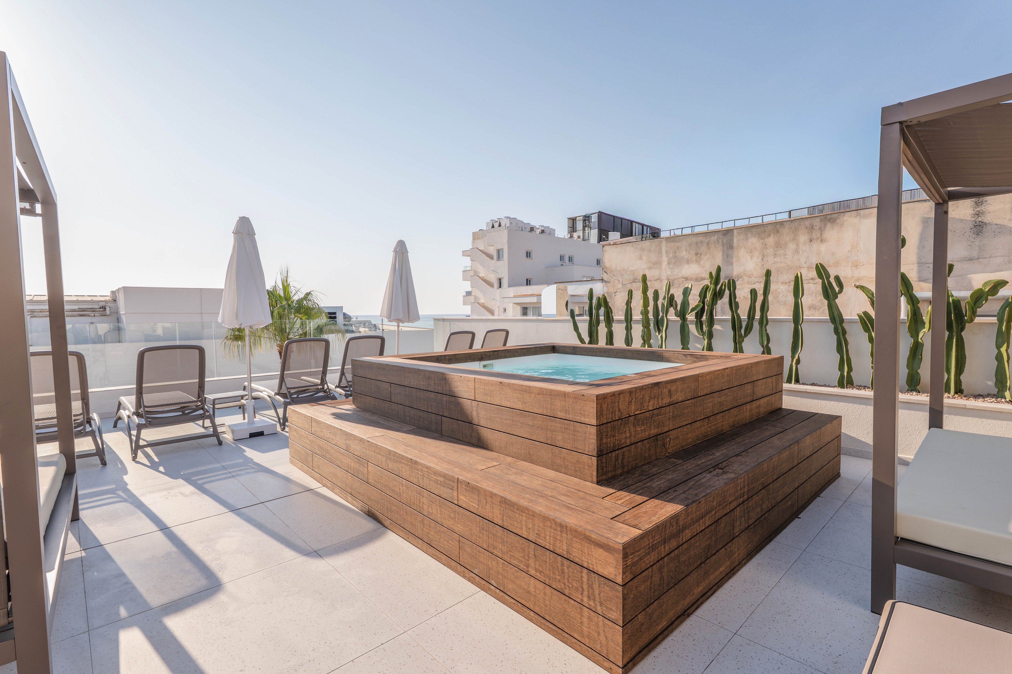 A contemporary rooftop terrace features a central wooden hot tub surrounded by sun loungers and large white umbrellas. The setting is bright and open, with clear skies and urban buildings in the background. Tall cacti line the wall, adding a touch of greenery to the minimalist, sunlit space.