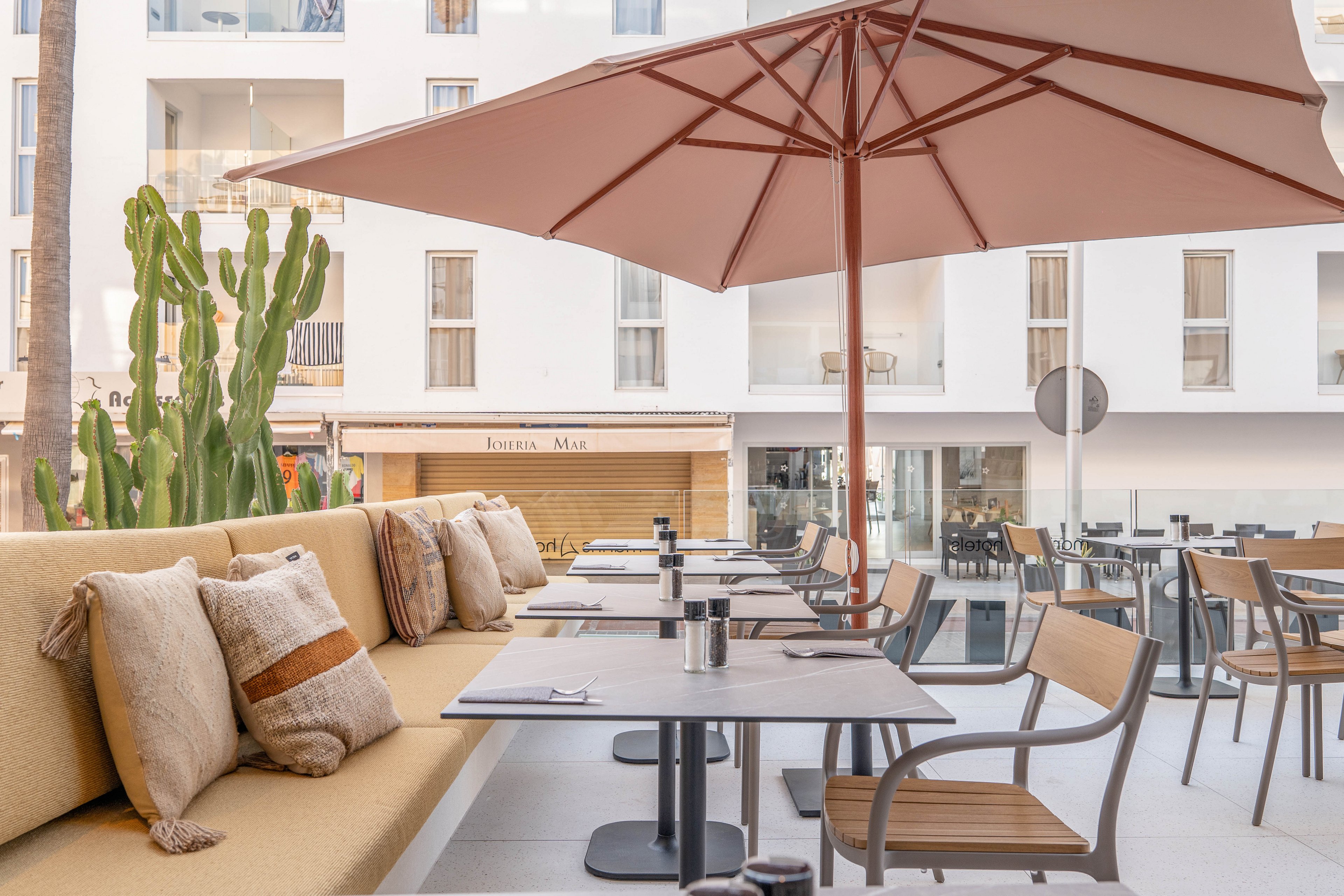 A stylish outdoor café terrace features cushioned bench seating with decorative pillows and wooden chairs around tables. A large umbrella provides shade, while a tall cactus adds a touch of greenery. The setting is bright and modern, with a neutral color palette and a view of a contemporary building façade in the background. The space appears inviting and relaxed, ideal for casual dining or coffee.