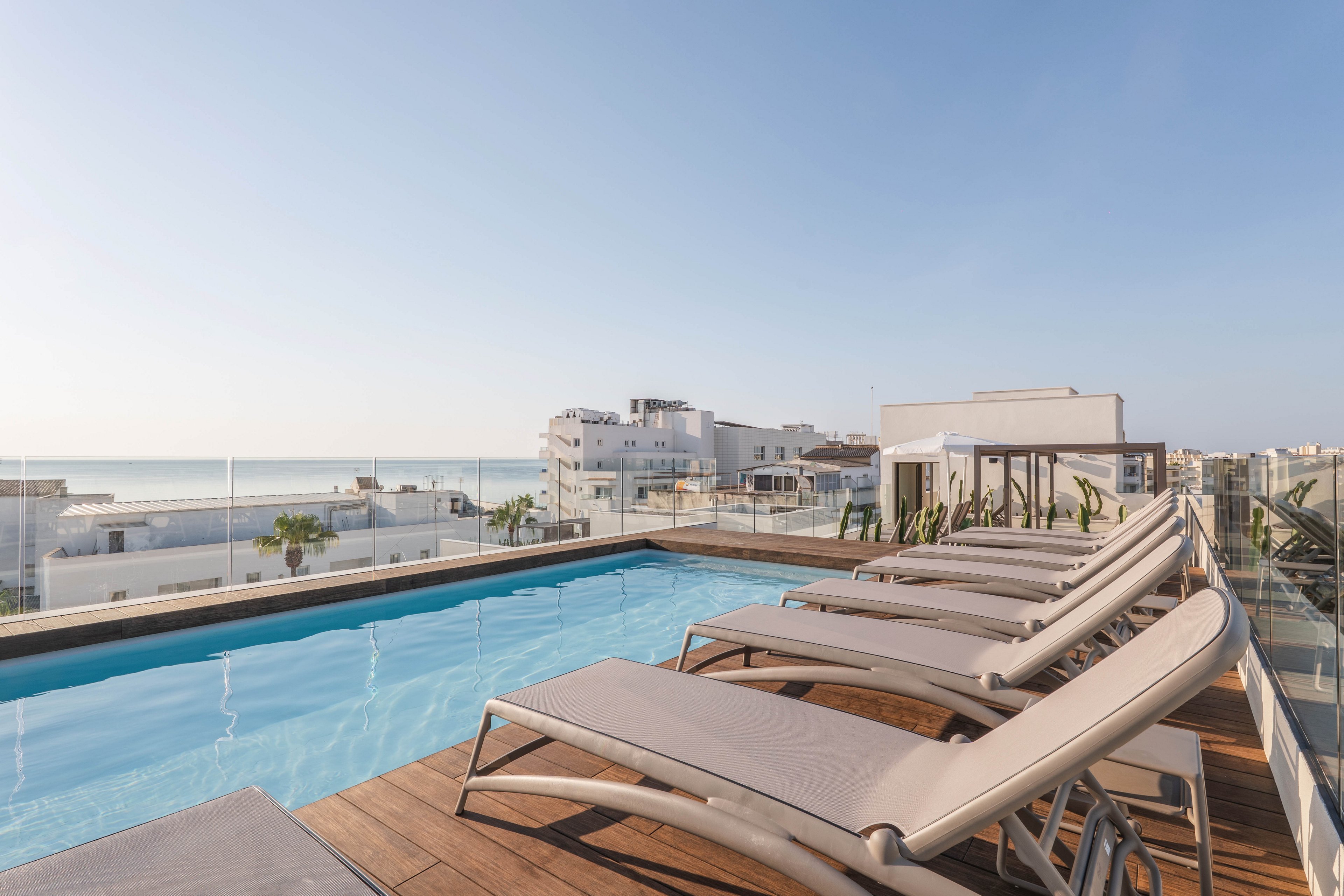 A contemporary rooftop swimming pool is surrounded by several sleek sun loungers arranged in a row. The setting overlooks a cityscape with white buildings and a distant view of the sea under a clear blue sky. Glass railings enhance the open, airy atmosphere, and the scene is bathed in natural daylight, creating a tranquil and inviting mood.