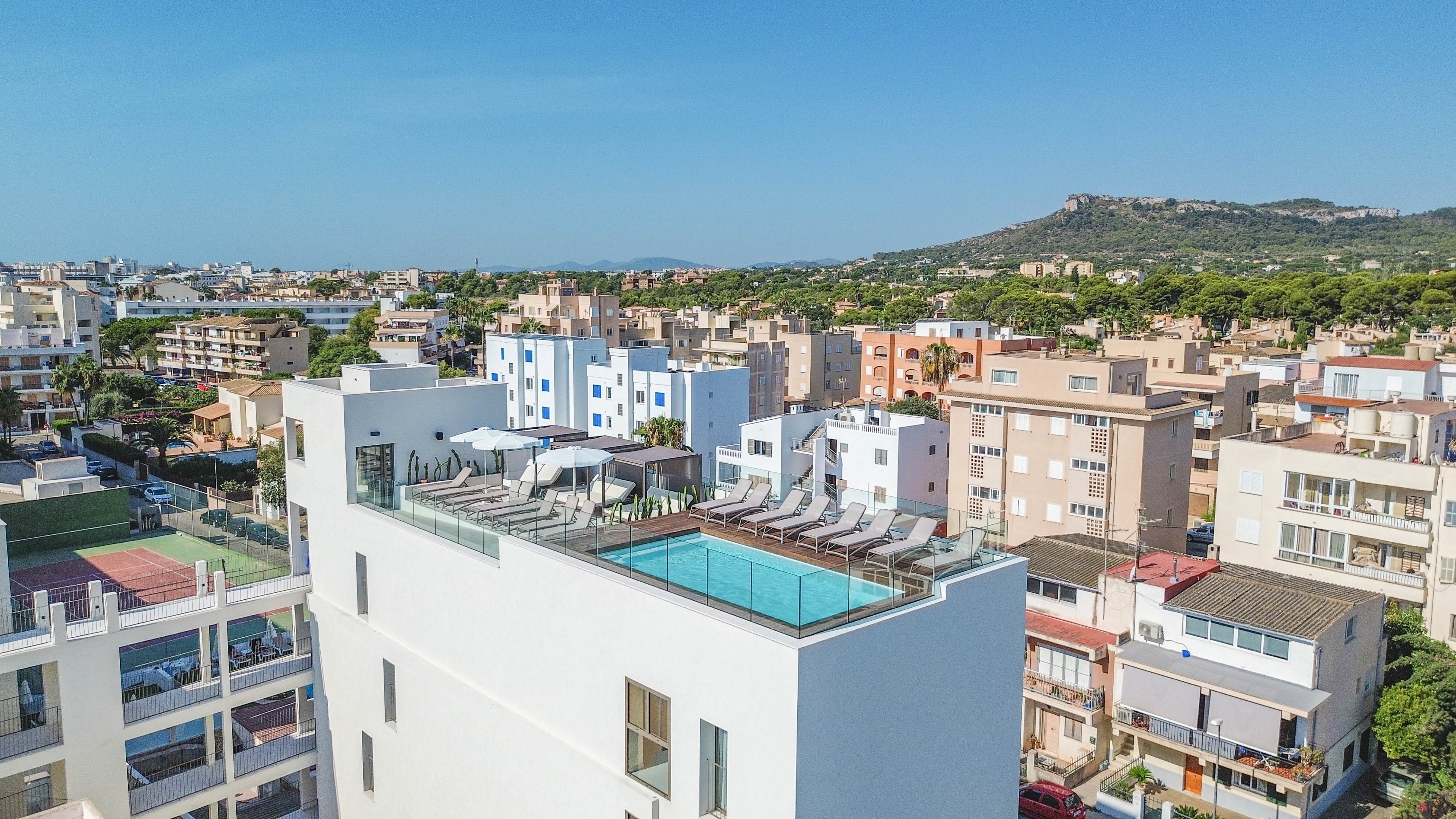 A contemporary white building features a rooftop swimming pool with lounge chairs and umbrellas, set against a backdrop of a sprawling city and distant green hills. The pool area is elevated, offering panoramic views of the surrounding urban landscape and natural scenery. The scene is bright and sunny, with clear blue skies and a relaxed, inviting atmosphere.