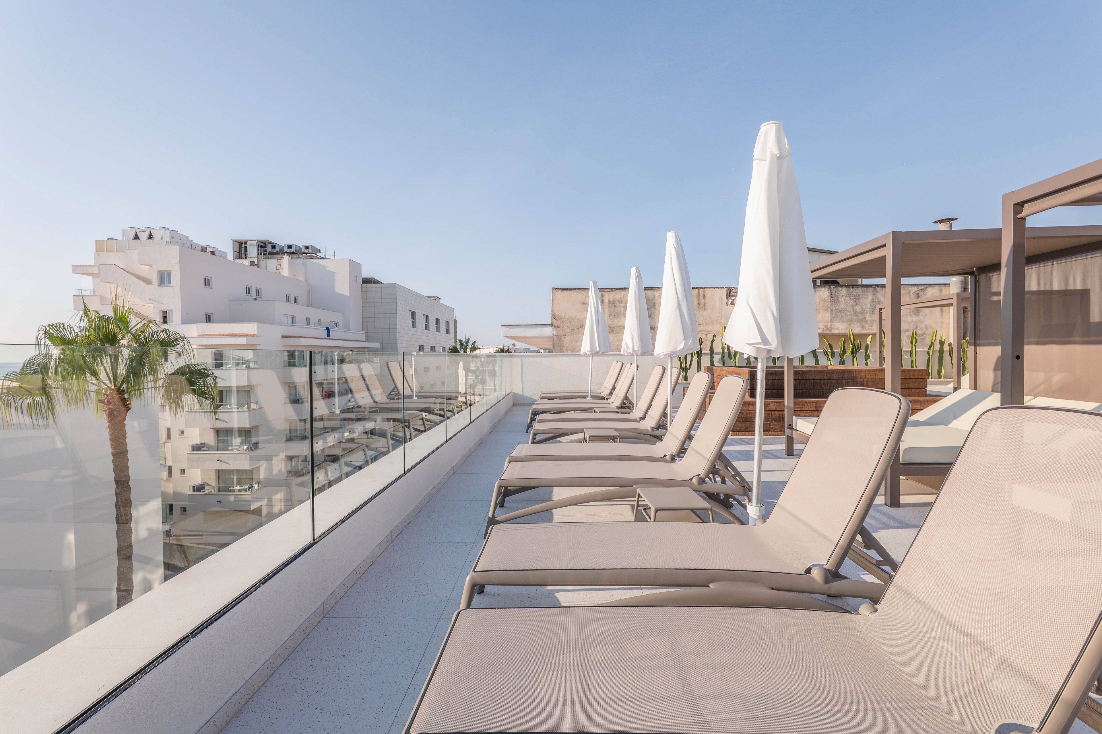 A row of beige sun loungers lines a contemporary rooftop terrace under clear blue skies. White umbrellas provide shade, and glass railings offer views of nearby modern buildings and palm trees. The setting is bright and minimalistic, evoking a relaxed, vacation-like atmosphere. No people are present, emphasizing tranquility and leisure.