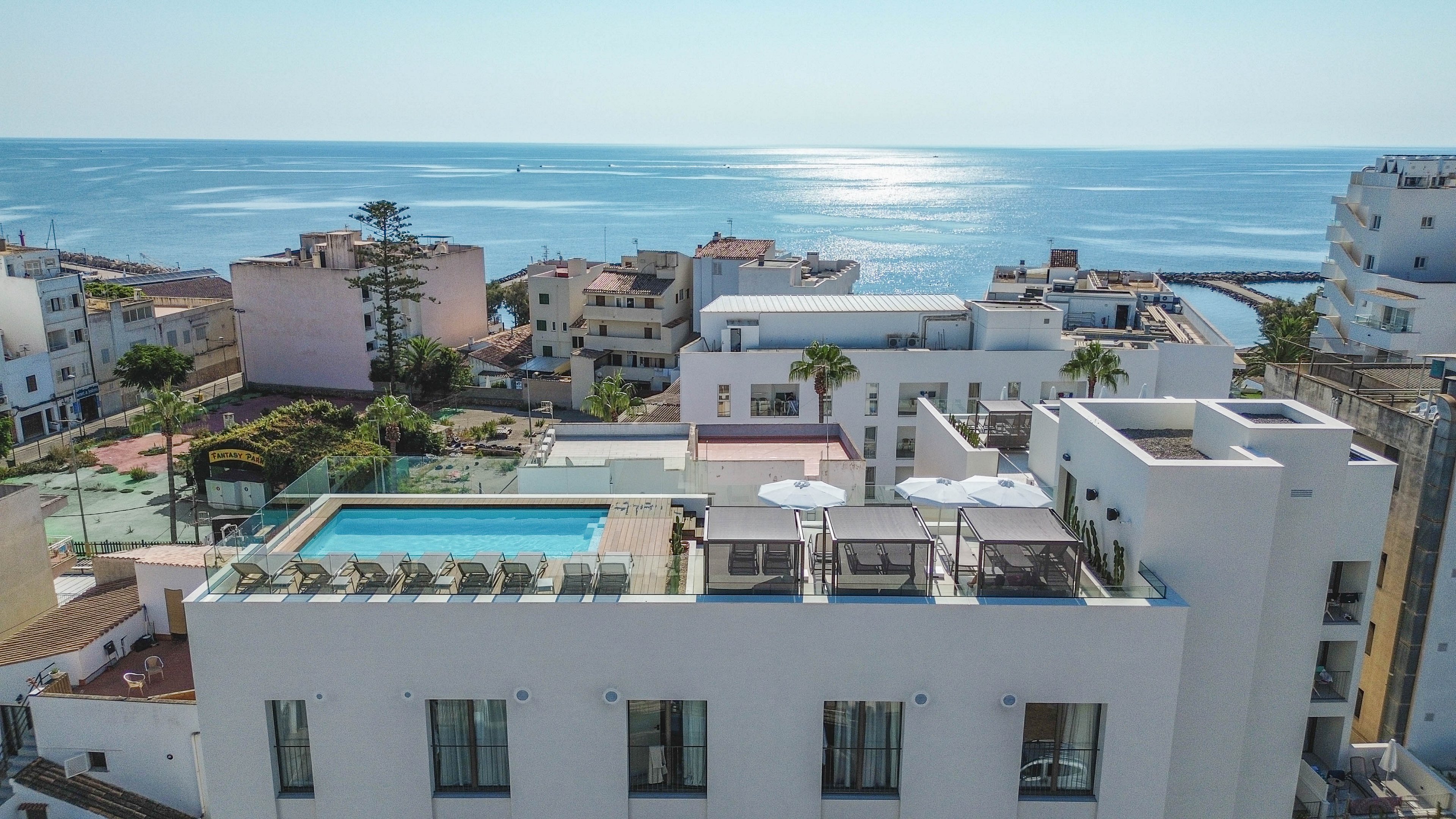 A contemporary white hotel building features a rooftop swimming pool overlooking the sea. The setting is a coastal urban area with several mid-rise buildings and a clear blue sky. Sun loungers and umbrellas are arranged around the pool, creating a relaxing atmosphere. The ocean glistens in the background, enhancing the tranquil, vacation-like mood.