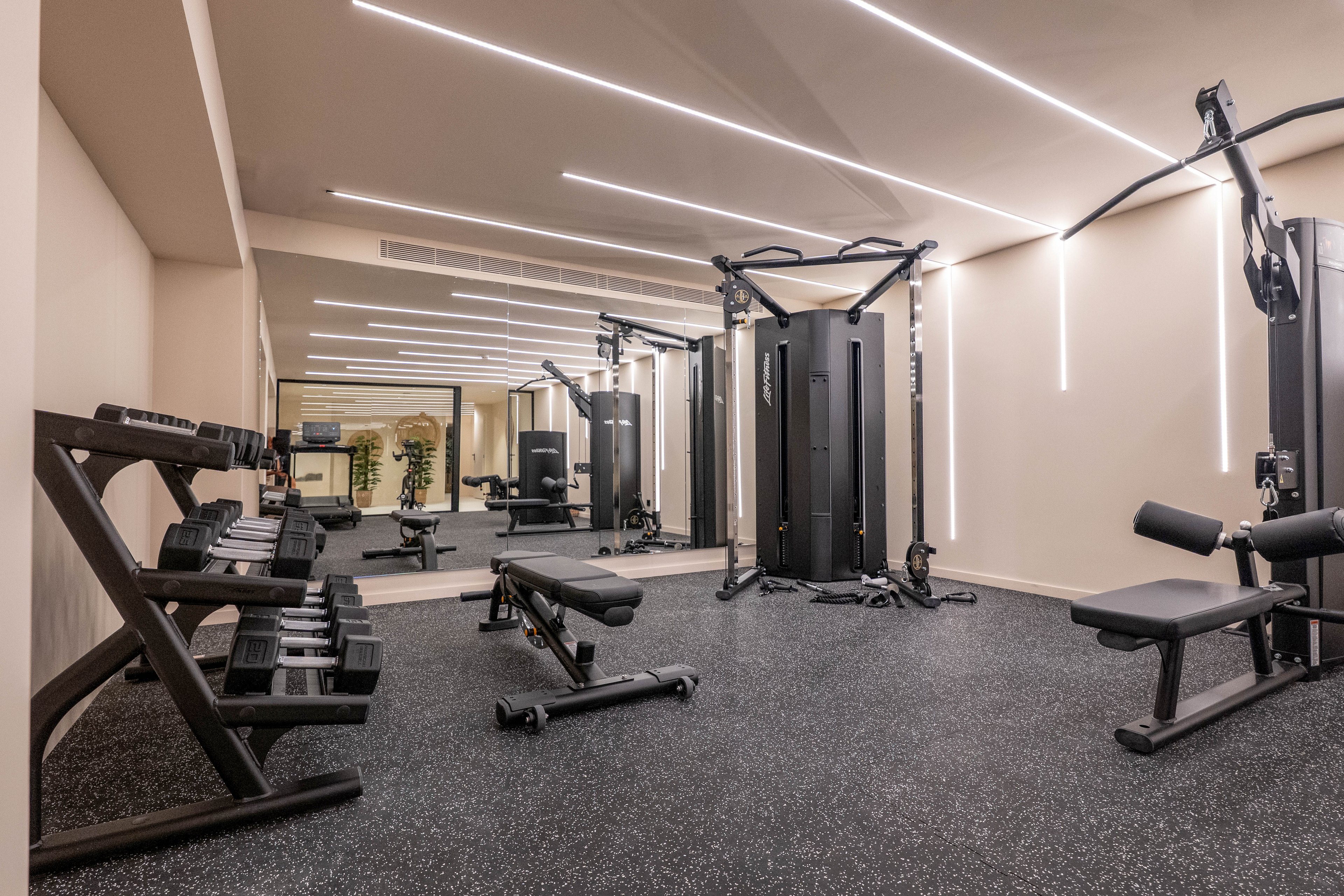 A spacious, contemporary gym features a variety of fitness equipment including free weights, benches, and multi-functional exercise machines. The room is well-lit with linear ceiling lights and has a large wall mirror that enhances the sense of space. The flooring is rubberized for safety and comfort, and the overall palette is neutral with black and gray tones. No people are present, emphasizing the clean and organized environment.