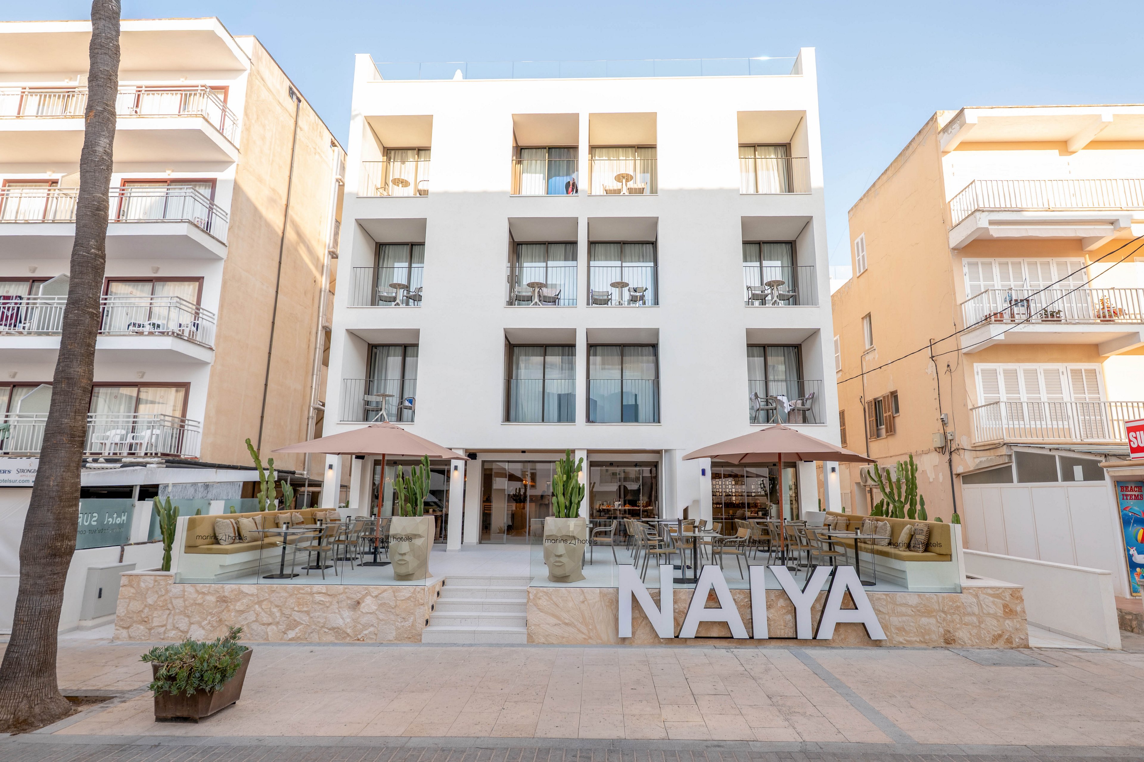 A contemporary white hotel building features balconies and a ground-level outdoor seating area. The setting is urban, with neighboring buildings and a wide pedestrian walkway in front. Large potted plants and umbrellas provide a welcoming atmosphere, while the prominent 'NAIYA' sign is clearly visible at the entrance.