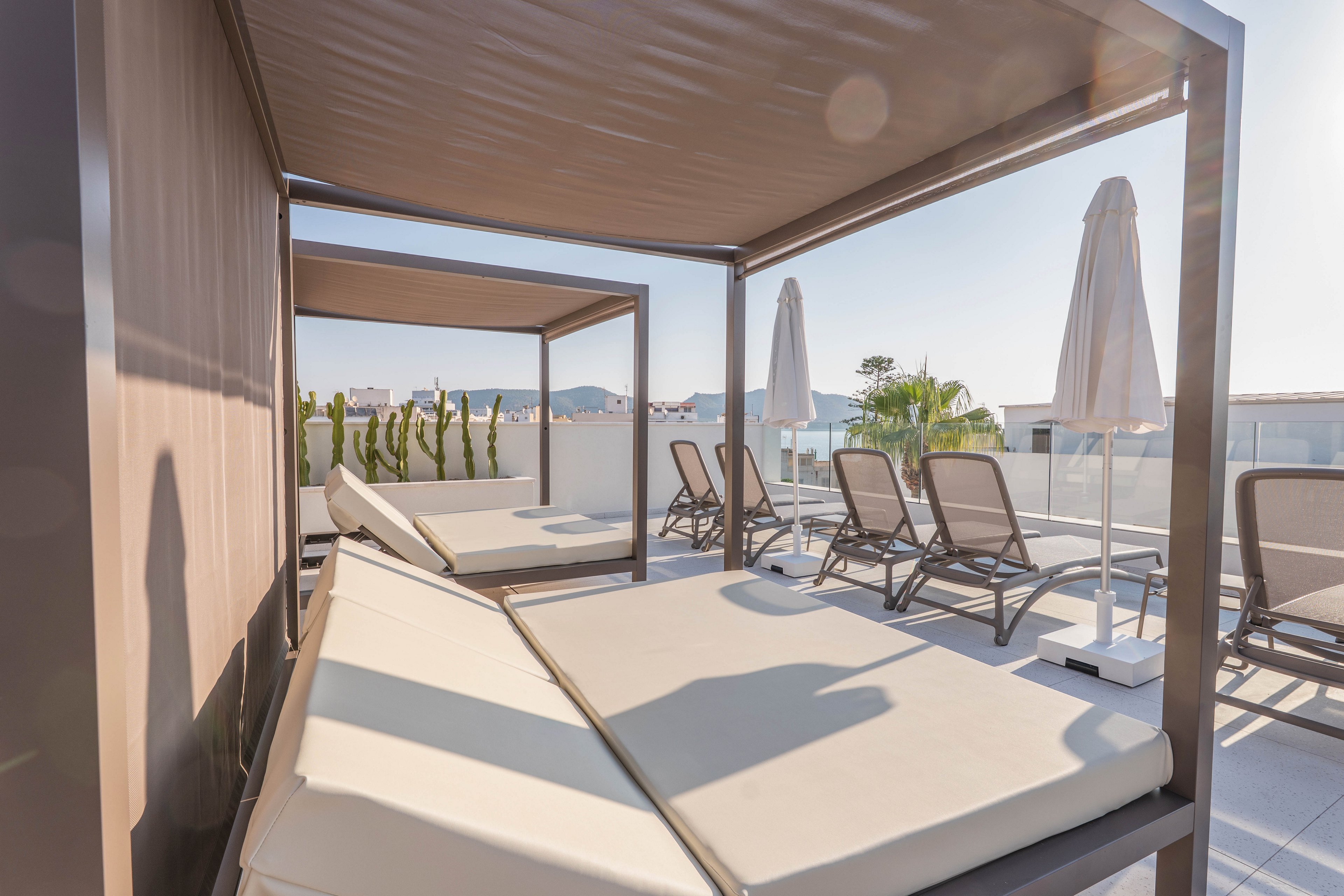 A contemporary rooftop terrace features large cushioned sunbeds under shaded canopies. Several reclining lounge chairs and white umbrellas are arranged on a tiled deck, with distant mountains and palm trees visible in the background. The scene is brightly lit with natural sunlight, creating a relaxed and inviting atmosphere.