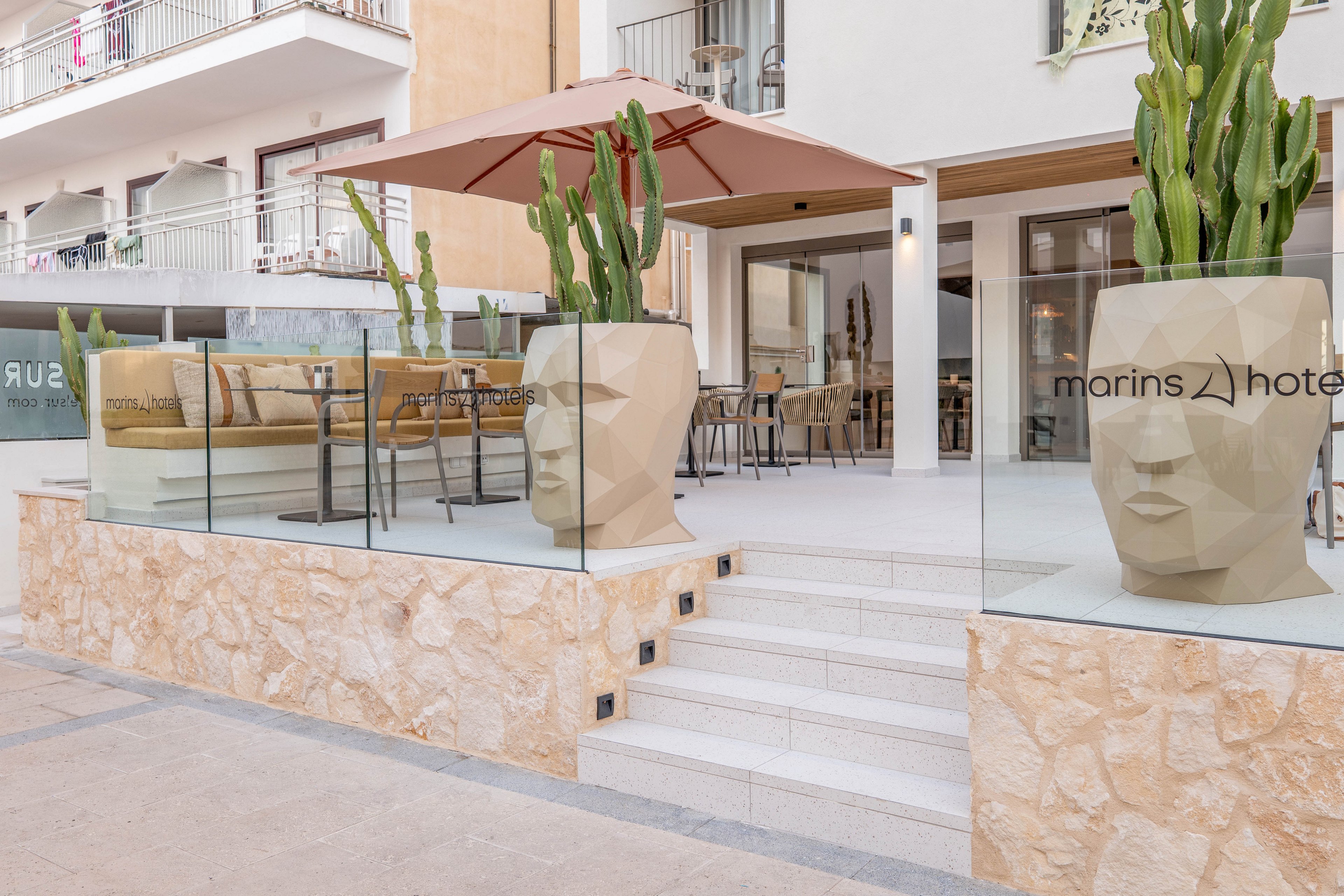 A contemporary hotel terrace features large geometric face-shaped planters with tall cacti, set against a backdrop of glass railings and beige stone walls. Outdoor seating with tables and chairs is arranged under a large umbrella, creating a welcoming atmosphere. The visible text 'marins hotel' appears on the glass, indicating the location. The setting is bright and airy, with a neutral color palette and minimalist design elements.