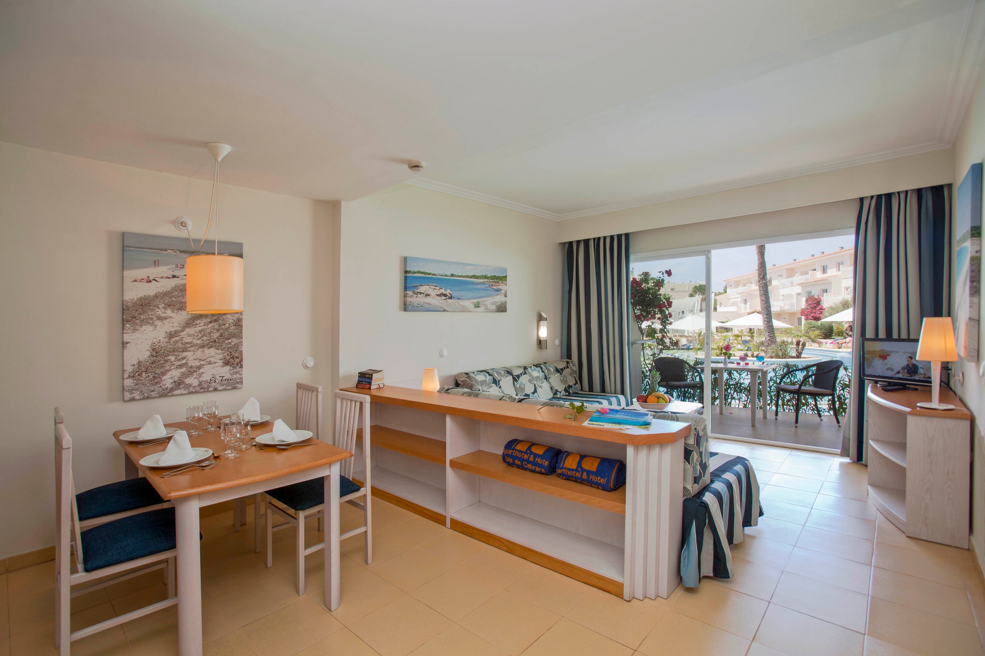 A spacious hotel suite features a small dining table set for four, a cozy living area with a sofa, and a balcony overlooking a sunny outdoor scene. The room is decorated in a coastal style with blue and beige tones, wall art, and soft lighting. Large glass doors allow natural light to fill the space, creating an inviting and relaxed atmosphere.