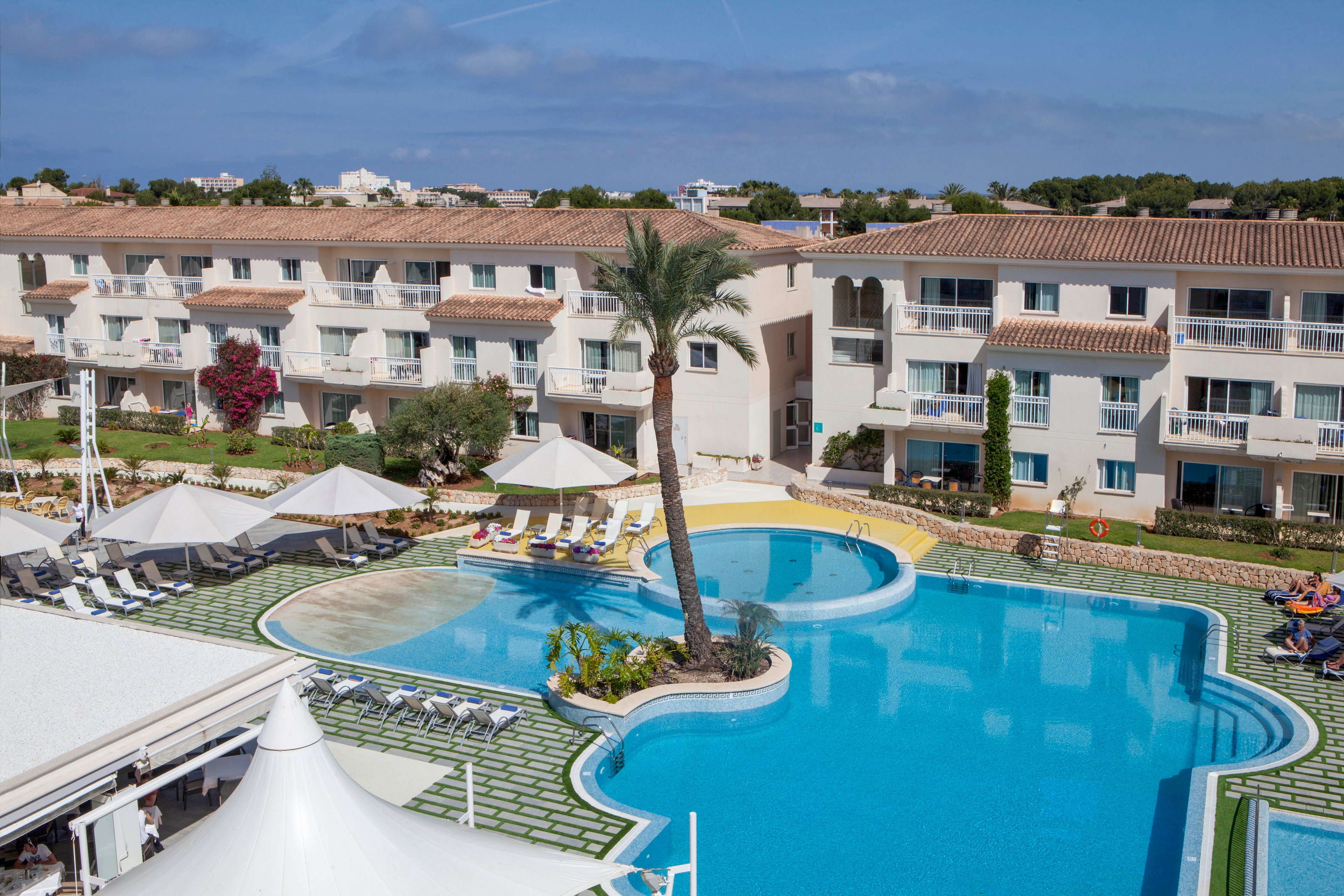 A spacious outdoor swimming pool is the central feature of this resort hotel, surrounded by sun loungers and umbrellas. The setting is bright and inviting, with well-maintained landscaping and a tall palm tree adding a tropical touch. The hotel buildings have a Mediterranean architectural style with balconies overlooking the pool area. The sky is clear, suggesting a warm and relaxing atmosphere ideal for vacationers.