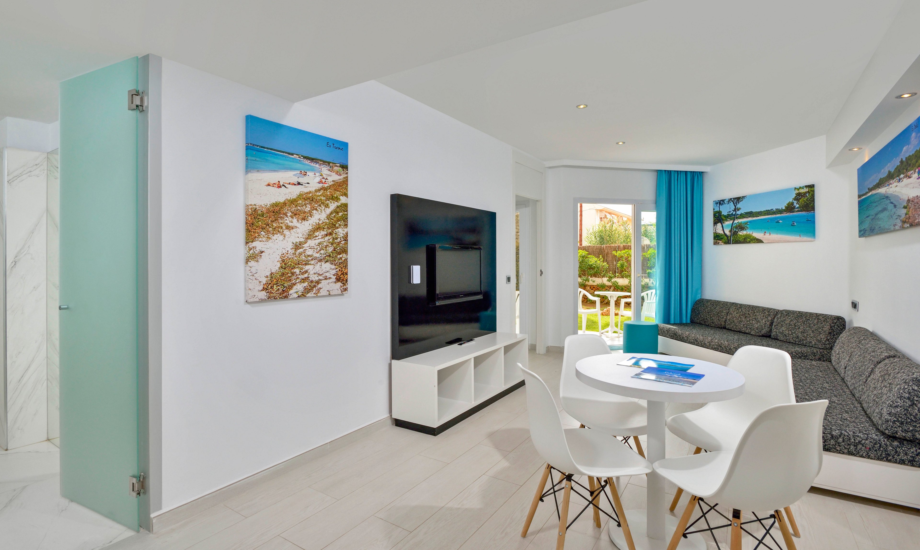 A contemporary living room features a large flat-screen TV mounted on a white entertainment unit, with a round dining table and four white chairs in the foreground. The space is decorated with vibrant beach landscape artwork and accented by turquoise curtains, creating a bright and inviting atmosphere. Natural light streams in from a glass door leading to a patio with greenery visible outside. The overall palette is clean and fresh, dominated by white and blue tones.