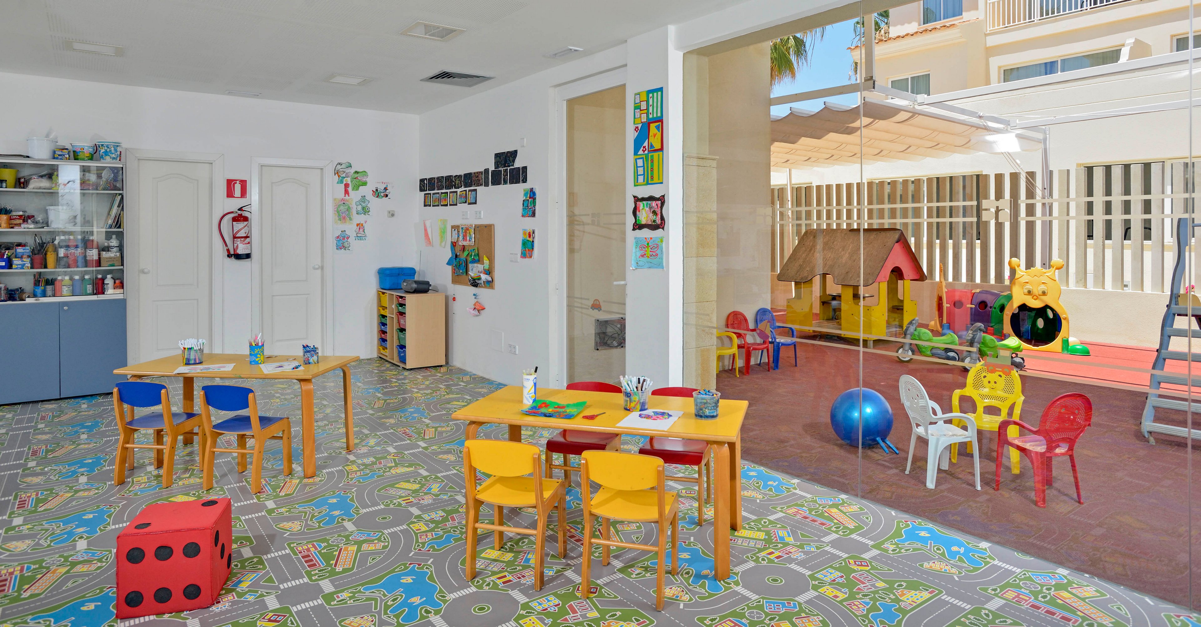 A colorful preschool classroom features small tables and chairs, art supplies, and playful wall decorations. The room opens to an outdoor play area with a playhouse, ride-on toys, and a large blue ball. The floor is covered with a patterned mat, and a large red foam die is visible. The setting is cheerful and organized, designed for young children’s activities and learning.