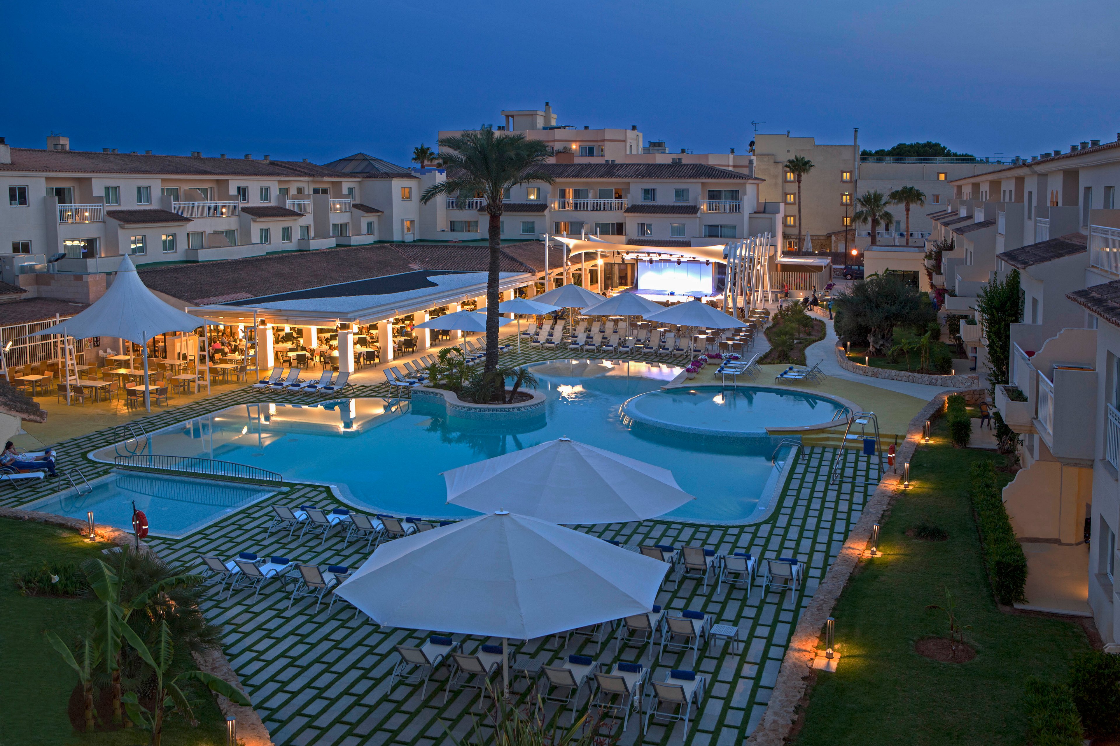 A large outdoor swimming pool is surrounded by lounge chairs and umbrellas at a resort complex during dusk. The scene is illuminated by soft lighting, creating a relaxed and inviting atmosphere. The resort buildings frame the pool, and a restaurant or bar area is visible with guests inside. The overall palette features cool blues and warm yellow lights, enhancing the tranquil mood.