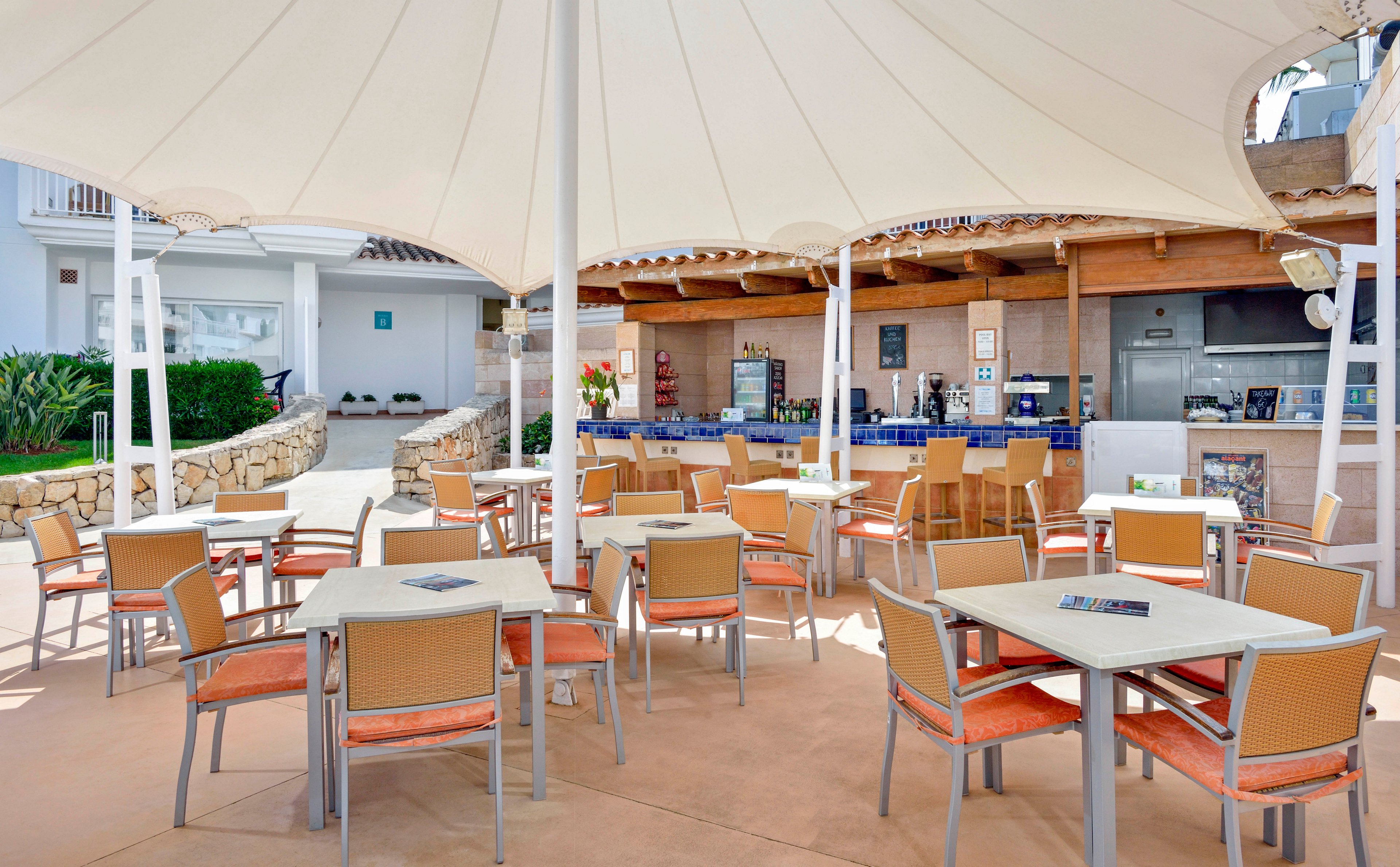 A modern outdoor cafe features multiple tables and chairs arranged beneath a large white canopy. The setting is bright and inviting, with a bar counter in the background and a stone wall lining the walkway. The overall mood is relaxed and summery, with neutral and orange tones dominating the palette. No people are present, and no visible text or numbers are seen in the image.