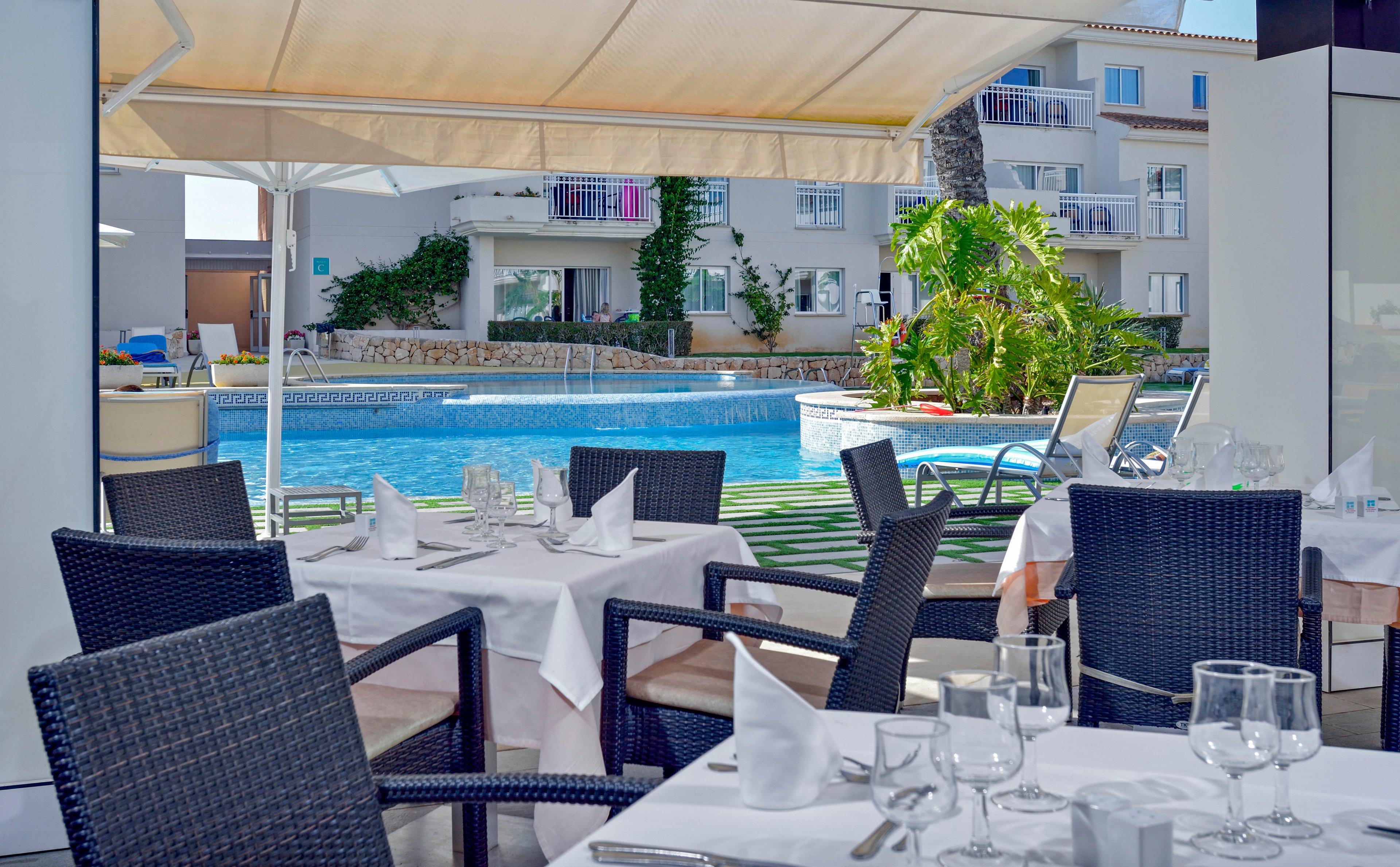 A neatly set outdoor restaurant table is positioned under a canopy, facing a clear blue swimming pool. The scene features black wicker chairs, white tablecloths, and glassware, with a resort-style apartment building in the background. The atmosphere is bright and inviting, with lush greenery and poolside lounge chairs visible. No people are present, emphasizing a tranquil and elegant setting. No visible text or numbers appear in the image.