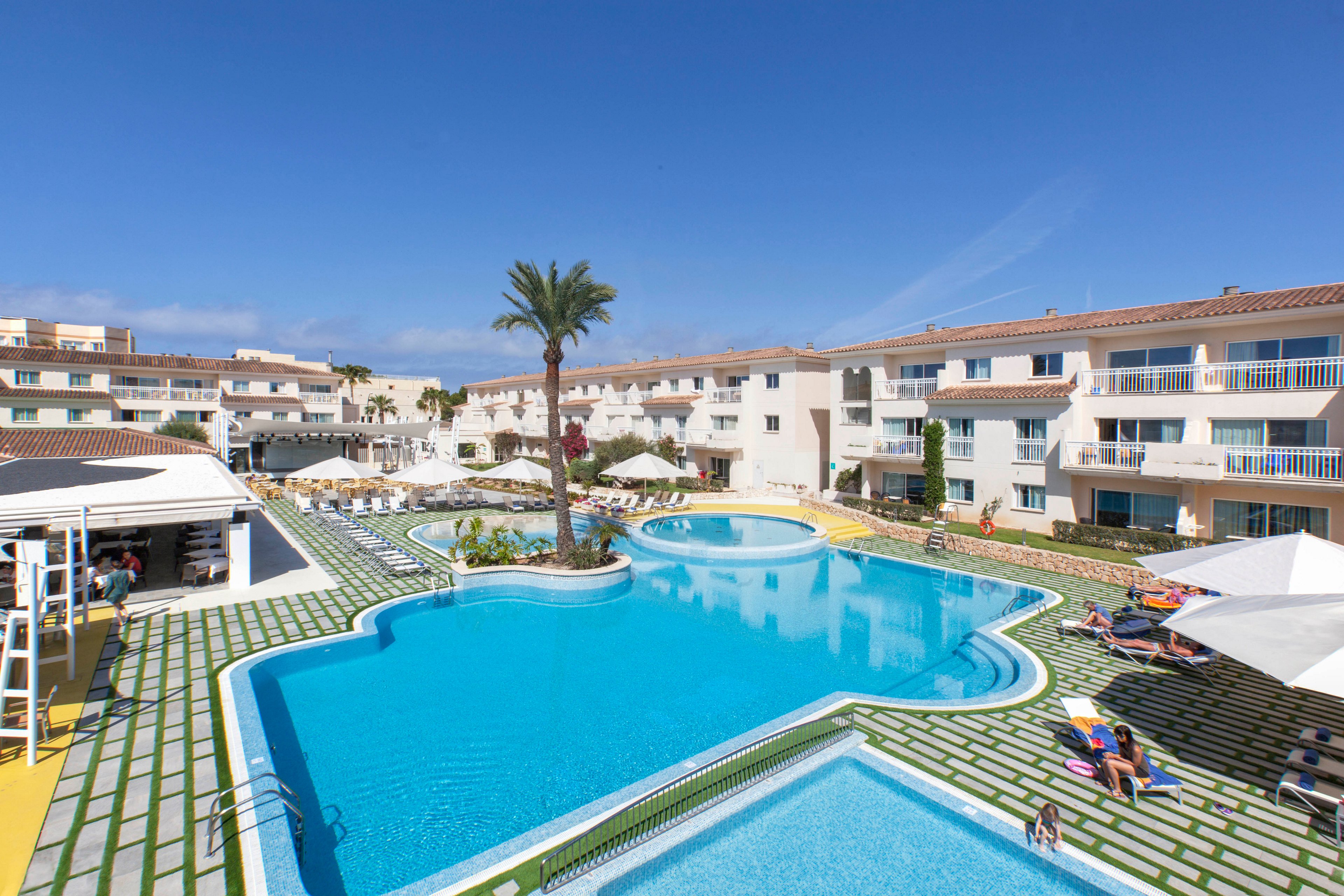 A large outdoor swimming pool is the central feature of a modern resort complex under a clear blue sky. The pool area is surrounded by white apartment-style buildings, sun loungers, and umbrellas, with a tall palm tree adding a tropical touch. Guests are relaxing by the poolside, enjoying the sunny weather and tranquil atmosphere. The setting suggests a vacation or leisure destination, with clean lines and a bright, inviting palette.