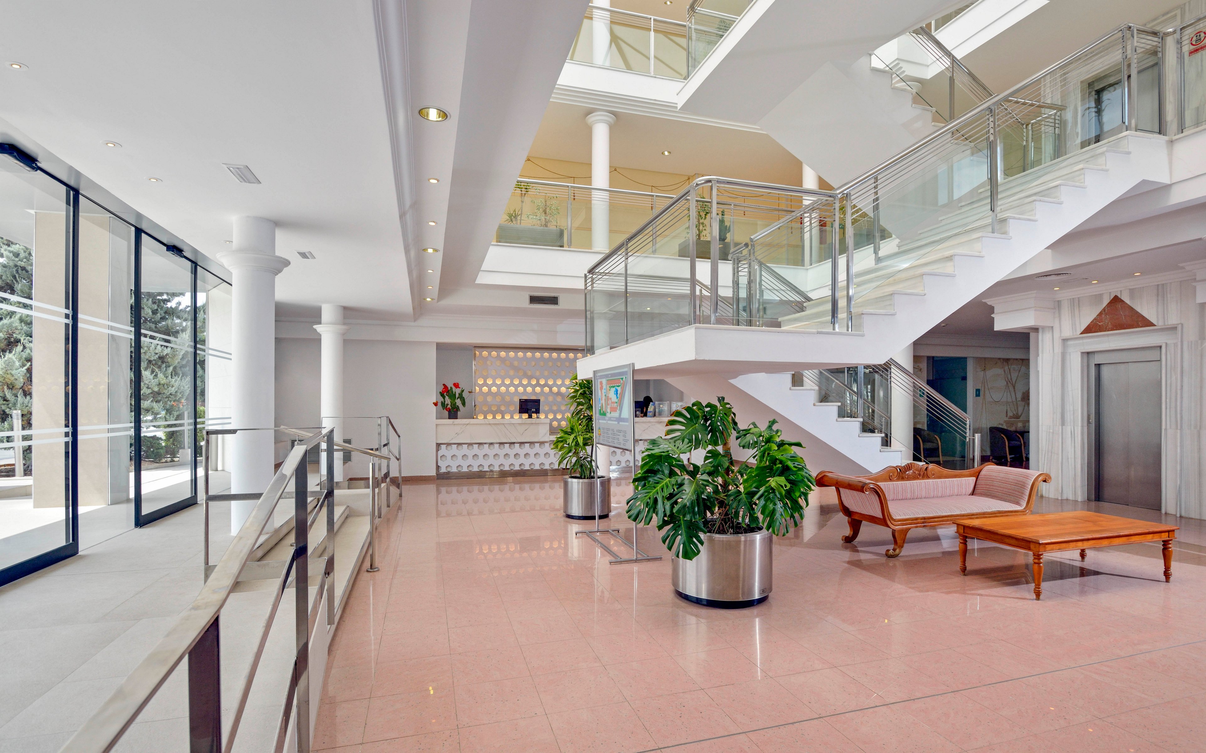 A spacious, contemporary hotel lobby features a large staircase with glass railings, polished pink floors, and a comfortable seating area with a wooden table and sofa. Potted green plants add a touch of nature, while large windows allow natural light to fill the space. The reception desk is visible in the background, and an elevator is located to the right.
