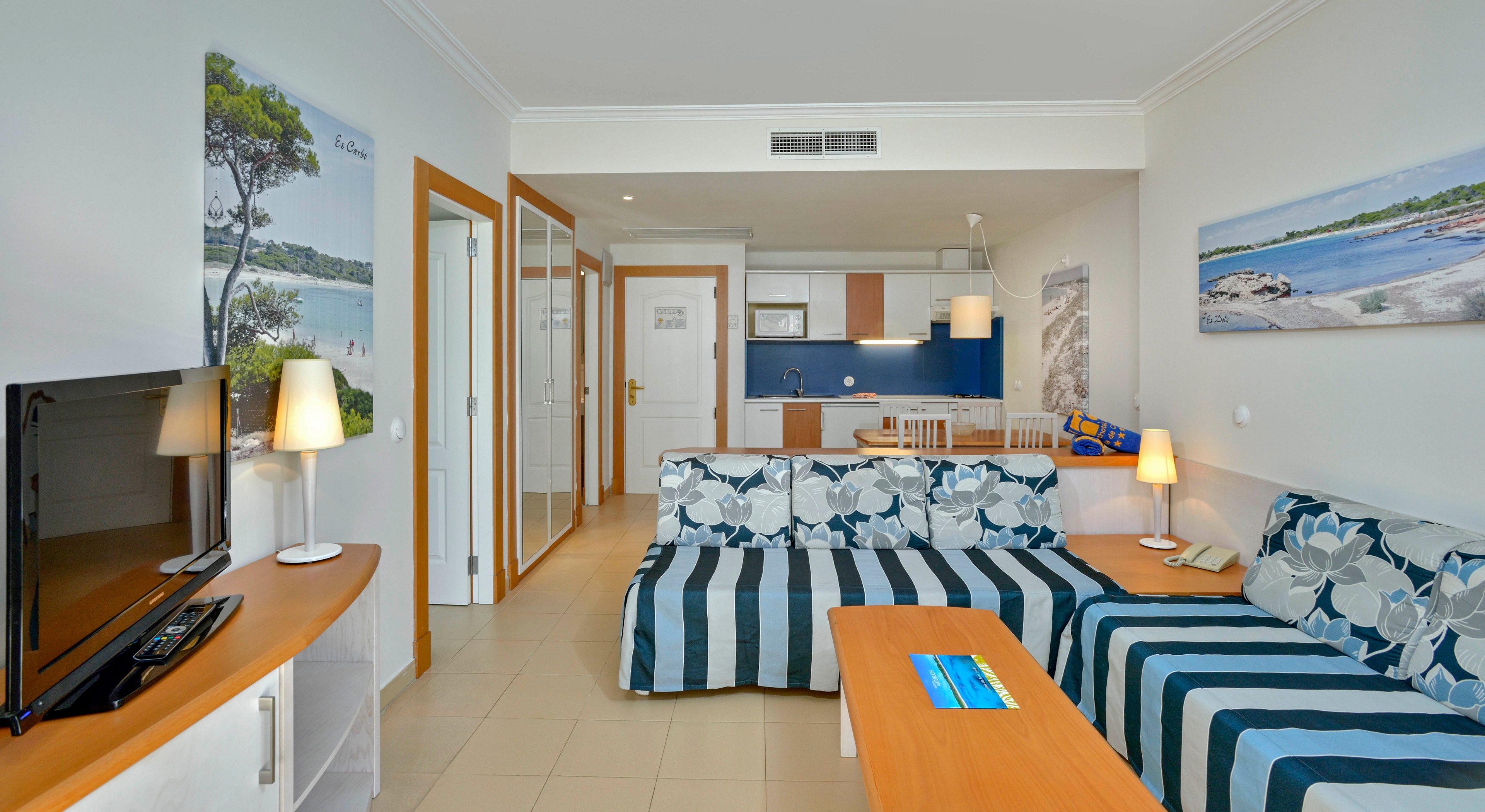 A bright, modern apartment living room features striped sofas, a flat-screen TV, and a wooden coffee table. The open-plan space includes a compact kitchen and dining area in the background, with wall art depicting coastal scenes. The room is well-lit with table lamps and natural light, creating a relaxed and inviting atmosphere.